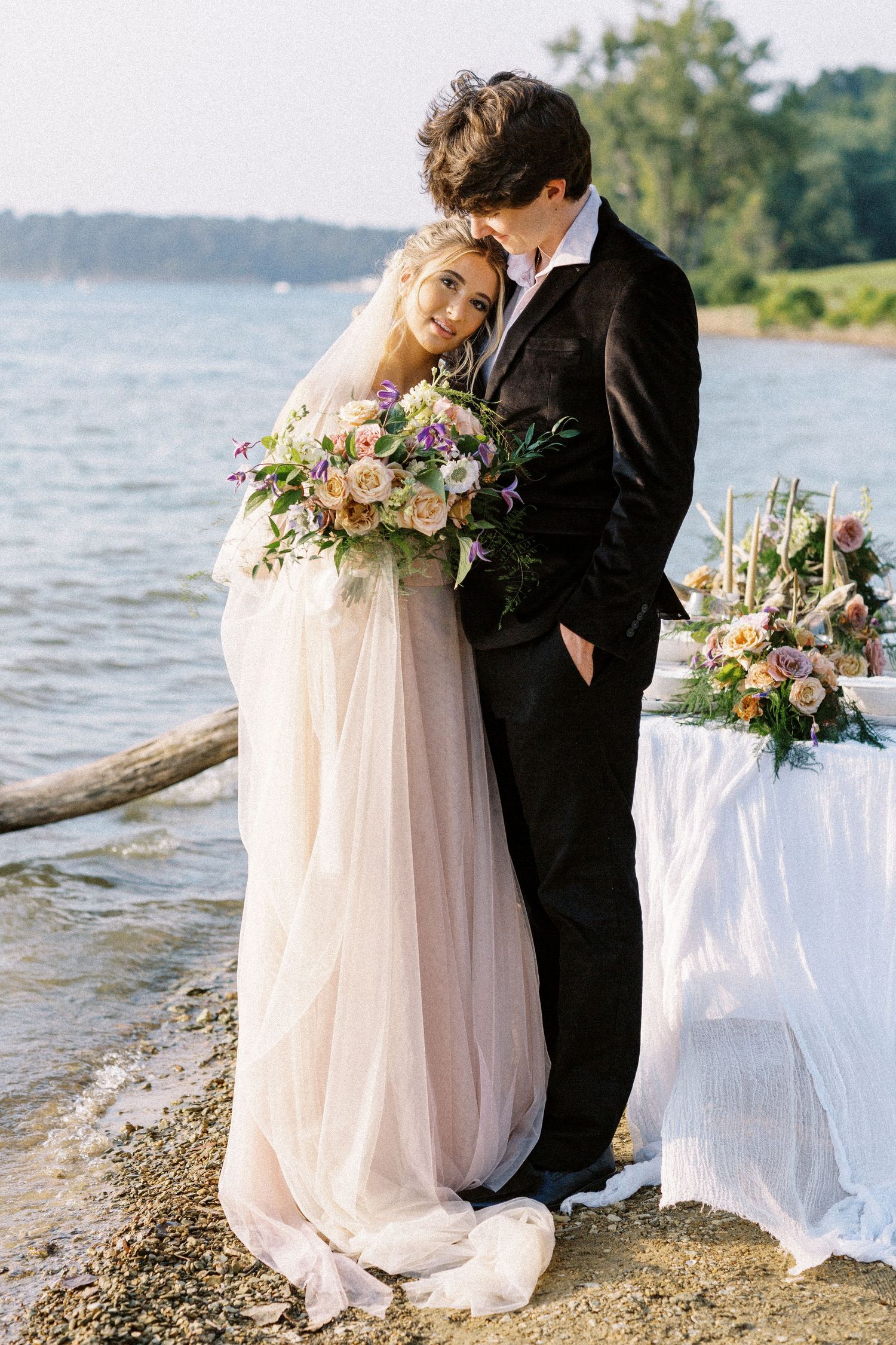 Romantic wedding couple embraces on beach shore with floral arrangements and white decor by waterfront.