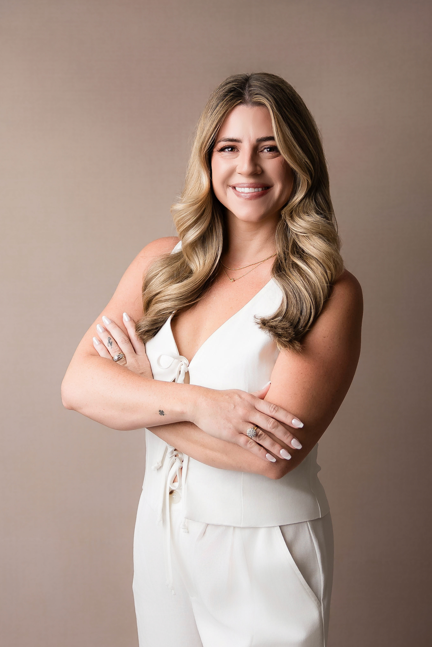 Professional portrait of a person with long blonde wavy hair wearing an elegant white outfit against a neutral background.