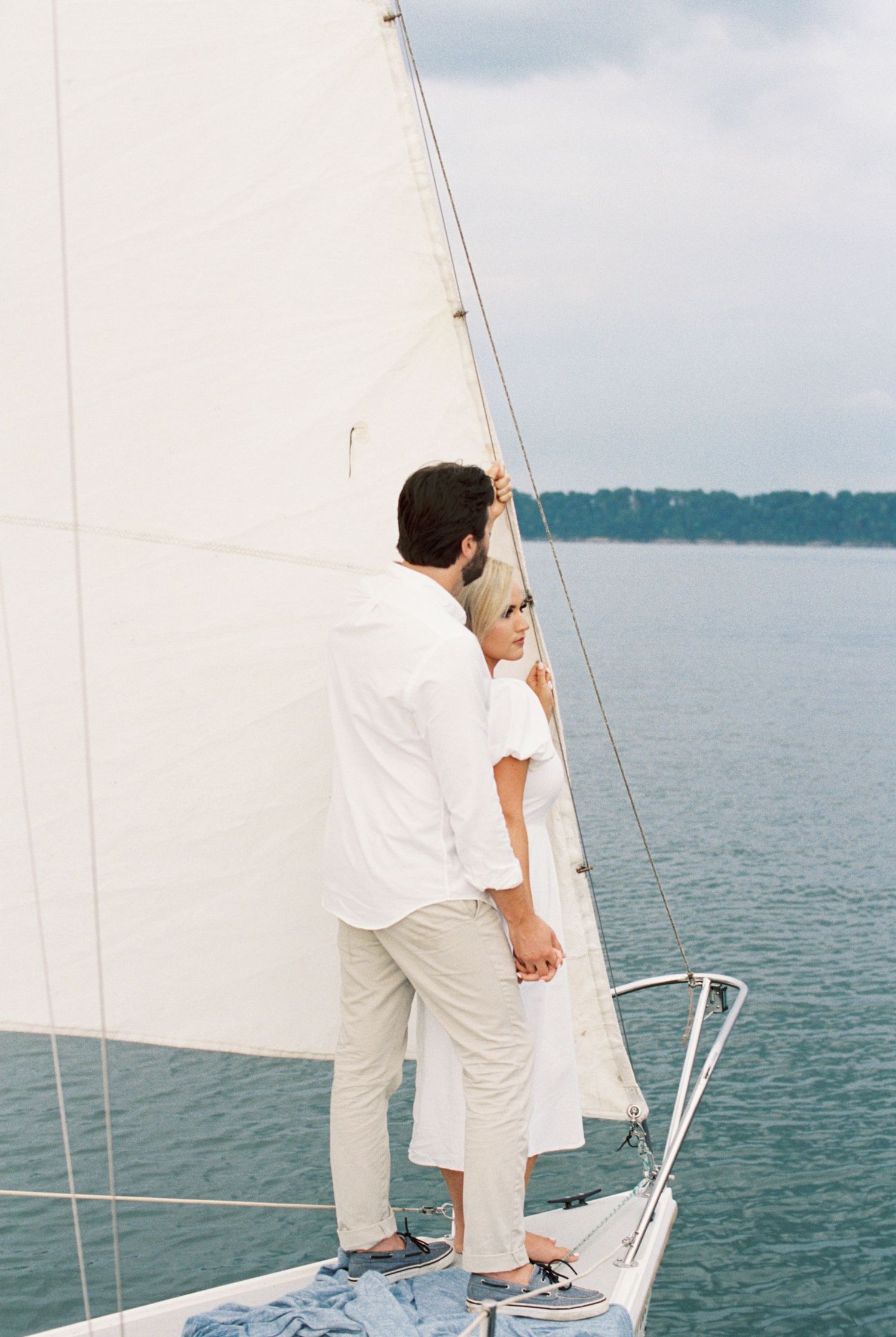 Two people in white outfits stand together on the bow of a sailboat on calm waters with white sails visible.