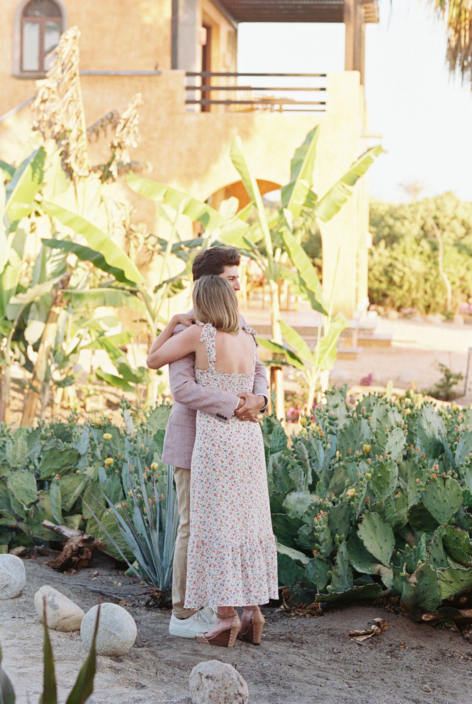 Two people share an embrace in a garden with cacti and tropical plants during golden hour outside a Mediterranean-style villa.