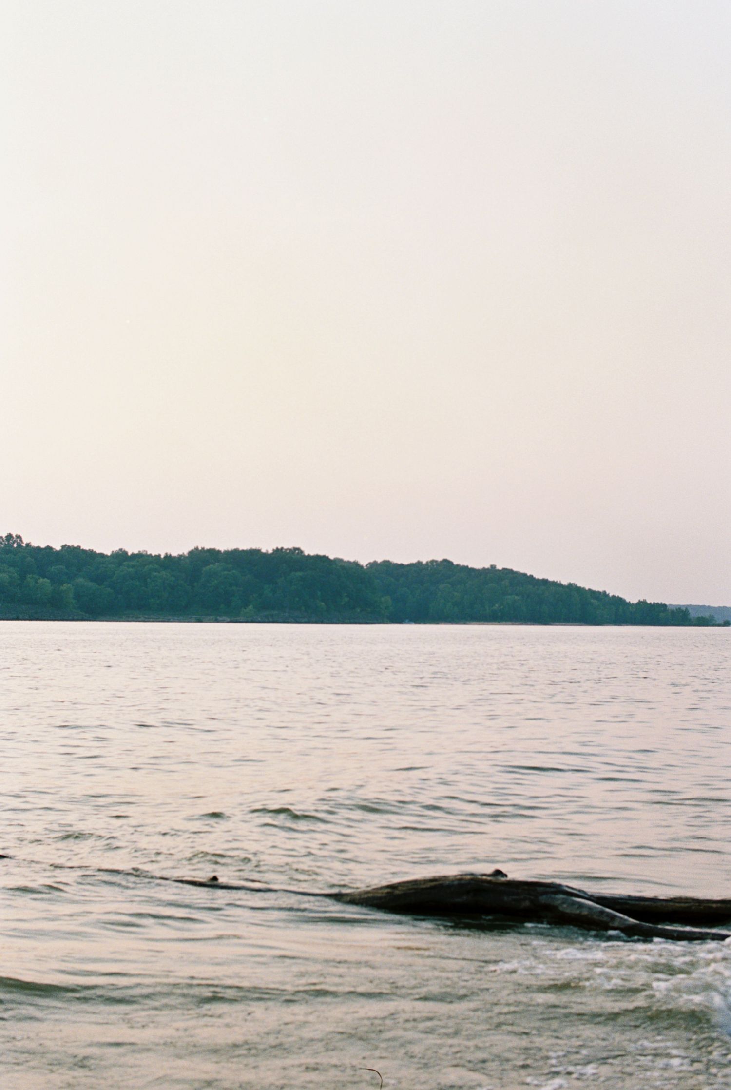 Peaceful lakeside scene with calm waters rippling against rocky shore and forested hills in the background.