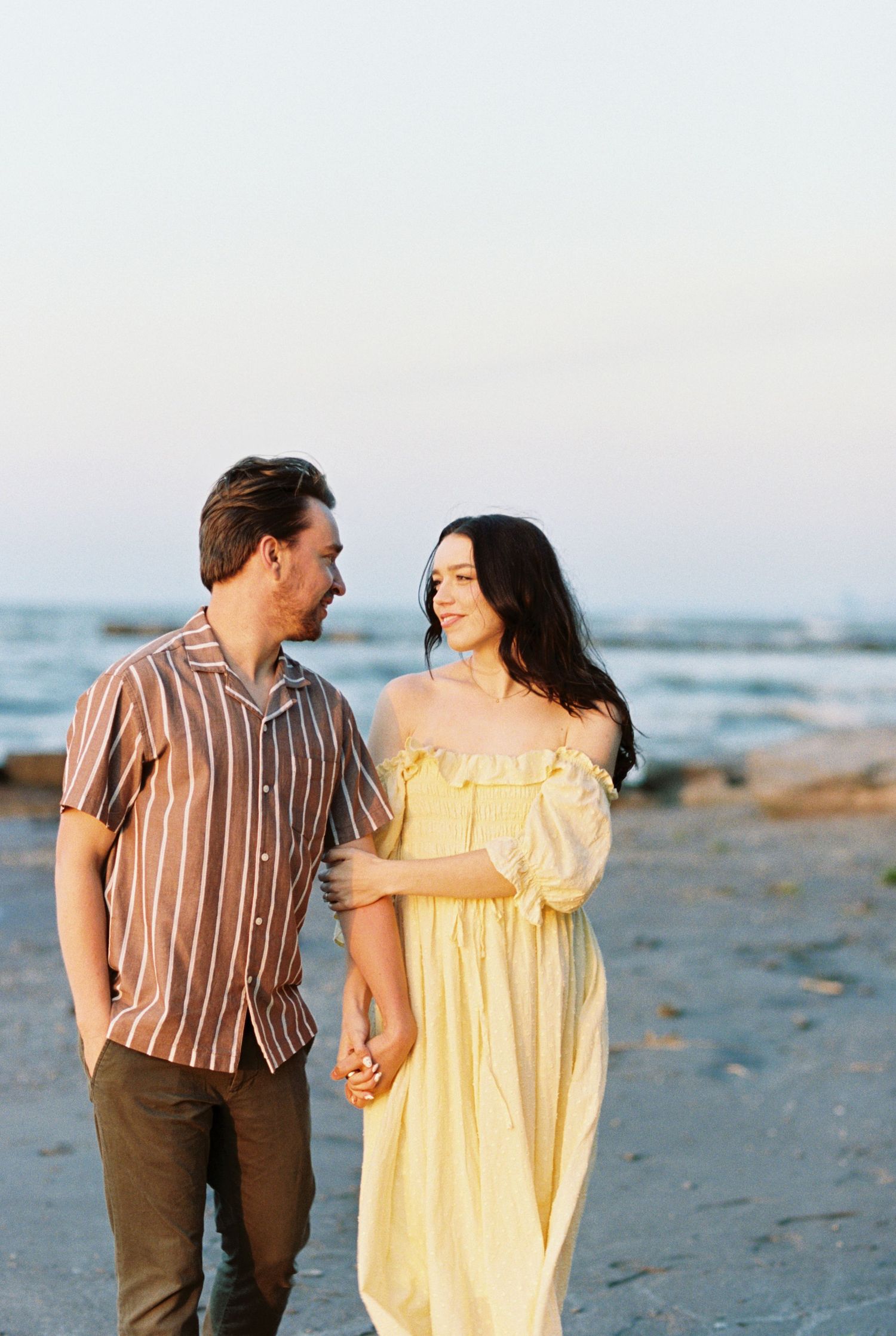 A couple holding hands while walking on a beach at sunset, with the woman wearing a yellow dress and man in striped shirt.