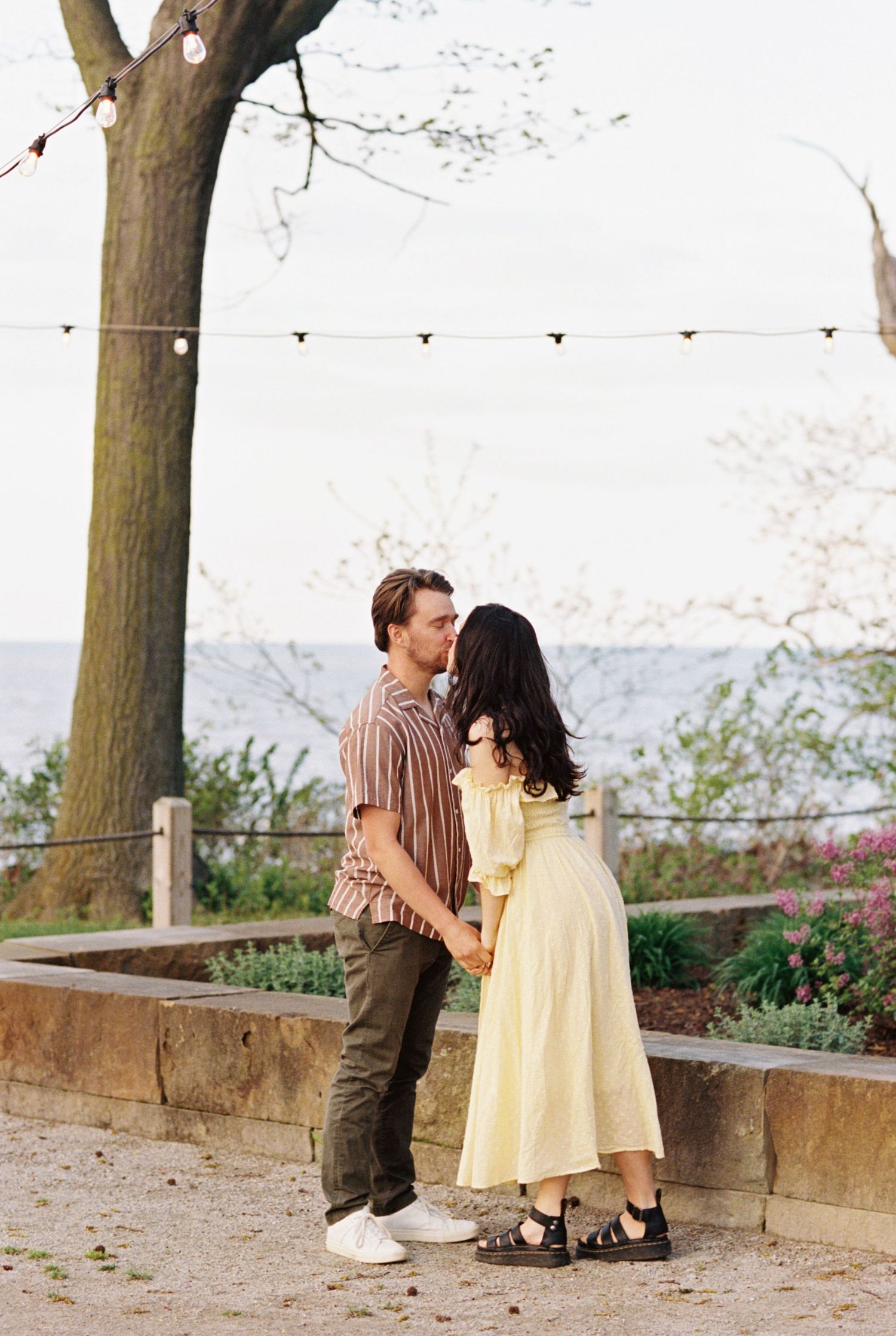 A romantic couple in casual attire share a tender moment on a scenic overlook with string lights overhead.