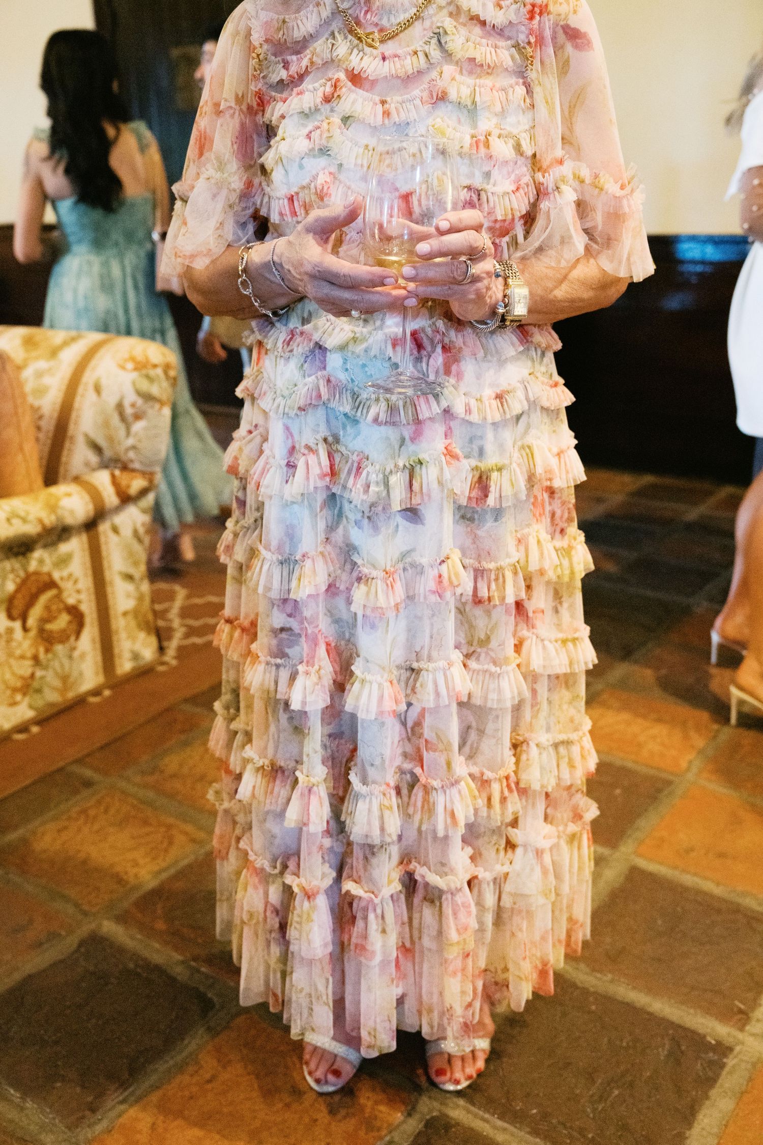 A romantic ruffled dress in soft pink and blue floral patterns features delicate tiers and sheer sleeves on a stone floor.