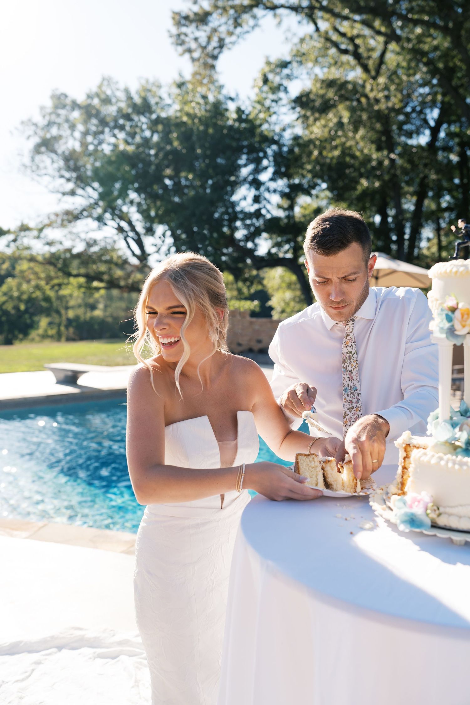 A couple in white attire cuts into an elegant wedding cake together by a swimming pool on a sunny day.