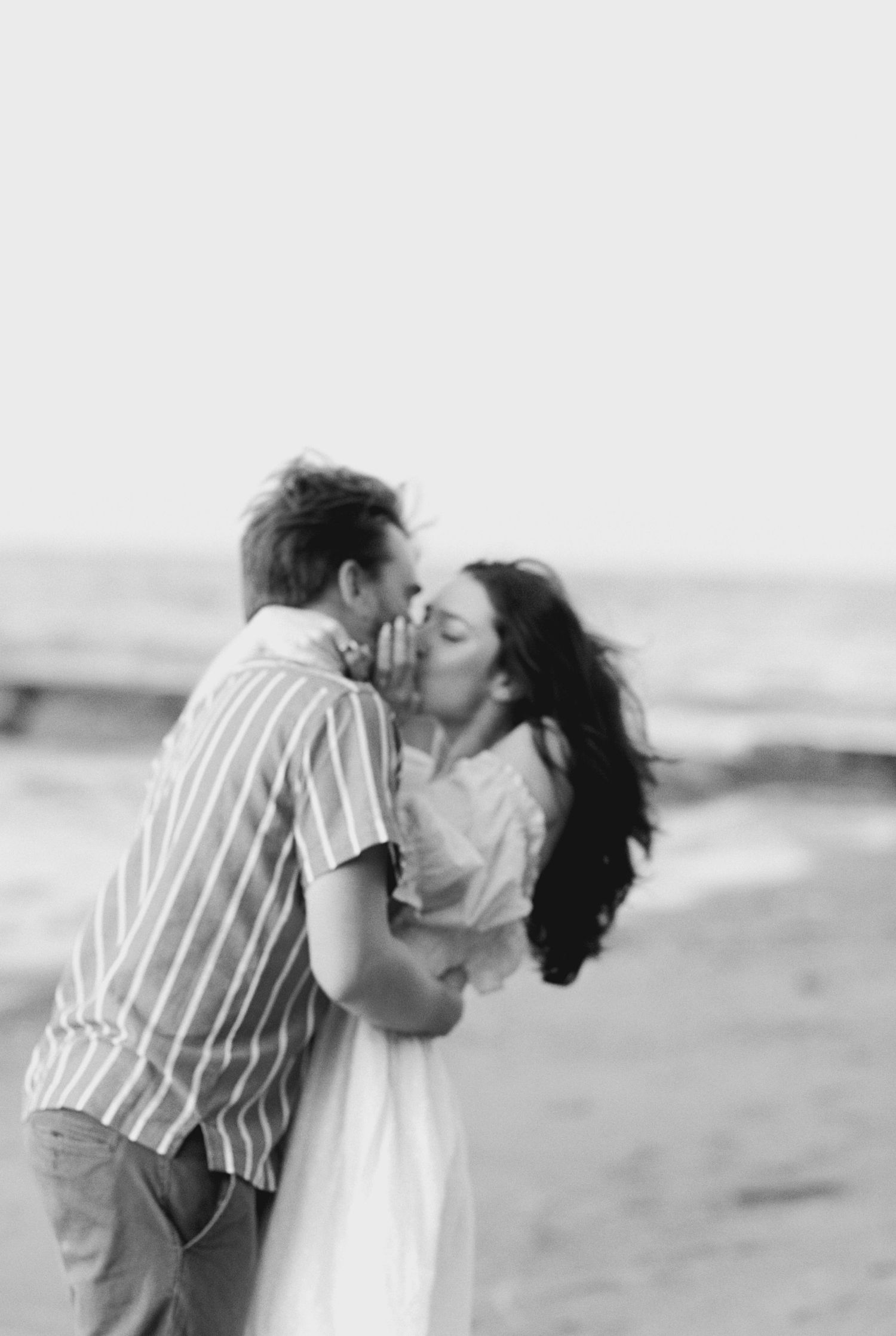 A romantic black and white photo of a couple sharing a tender kiss on the beach with ocean waves in the background.