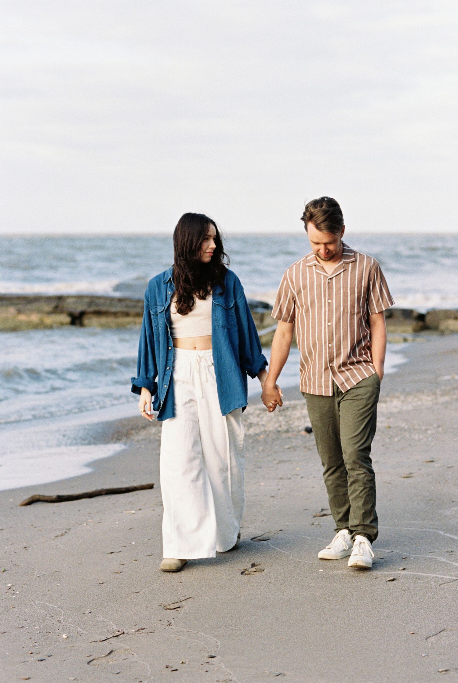 A couple walks hand in hand along a beach shoreline with ocean waves and cloudy sky in the background.