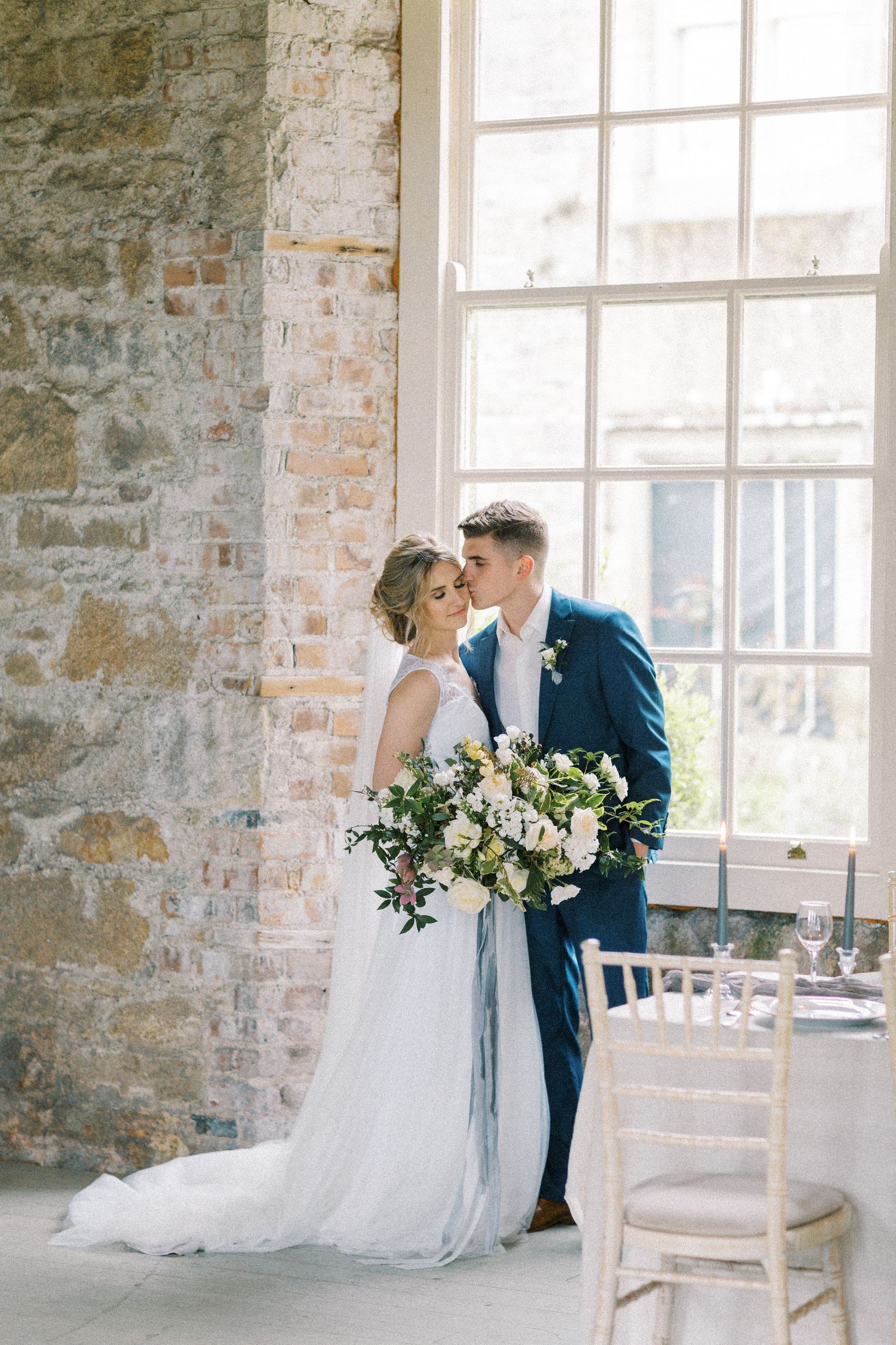 A romantic wedding couple stands by a rustic brick wall and window, the bride holding an elegant white and green bouquet.