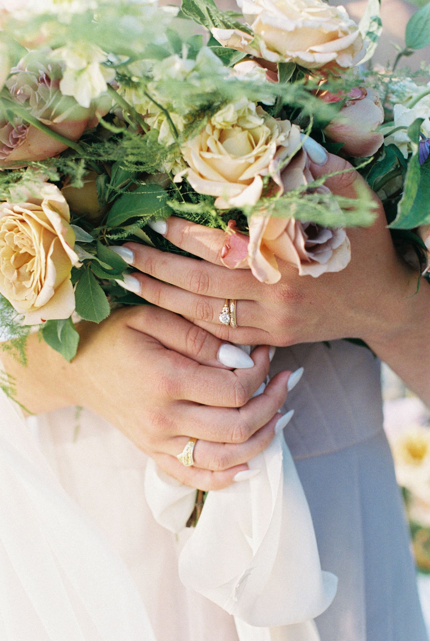 Close up of hands holding an elegant bouquet of cream roses with green foliage against a white wedding dress.