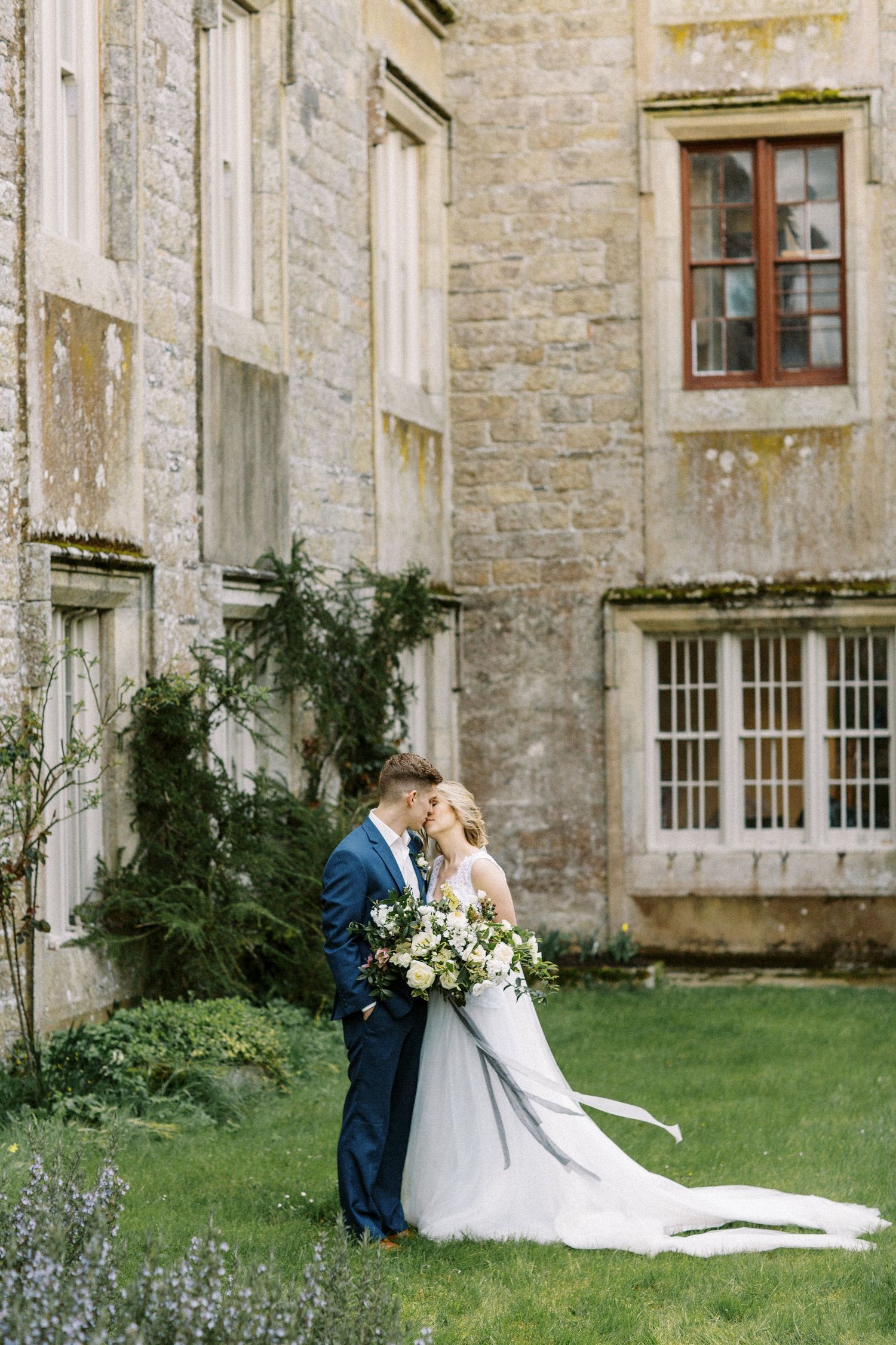 A couple in wedding attire share an intimate moment in front of a stone castle wall with climbing vines and windows.