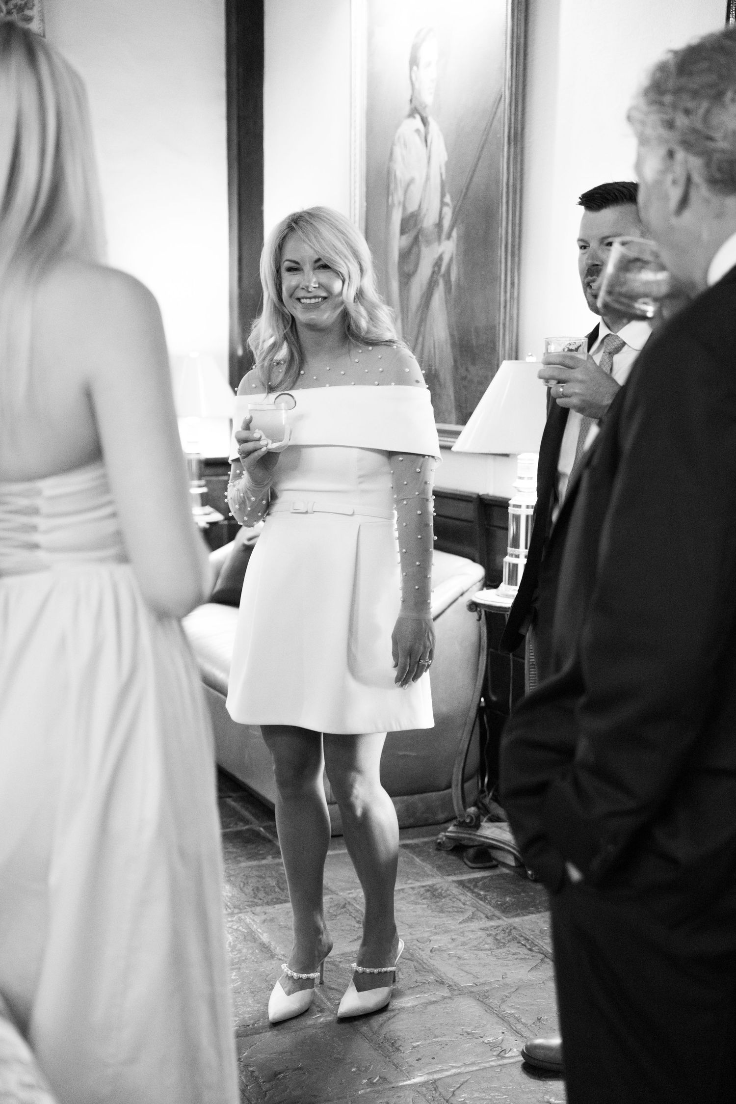 A celebration shows people gathered in a room with a person wearing an elegant white off-shoulder mini dress and flats.
