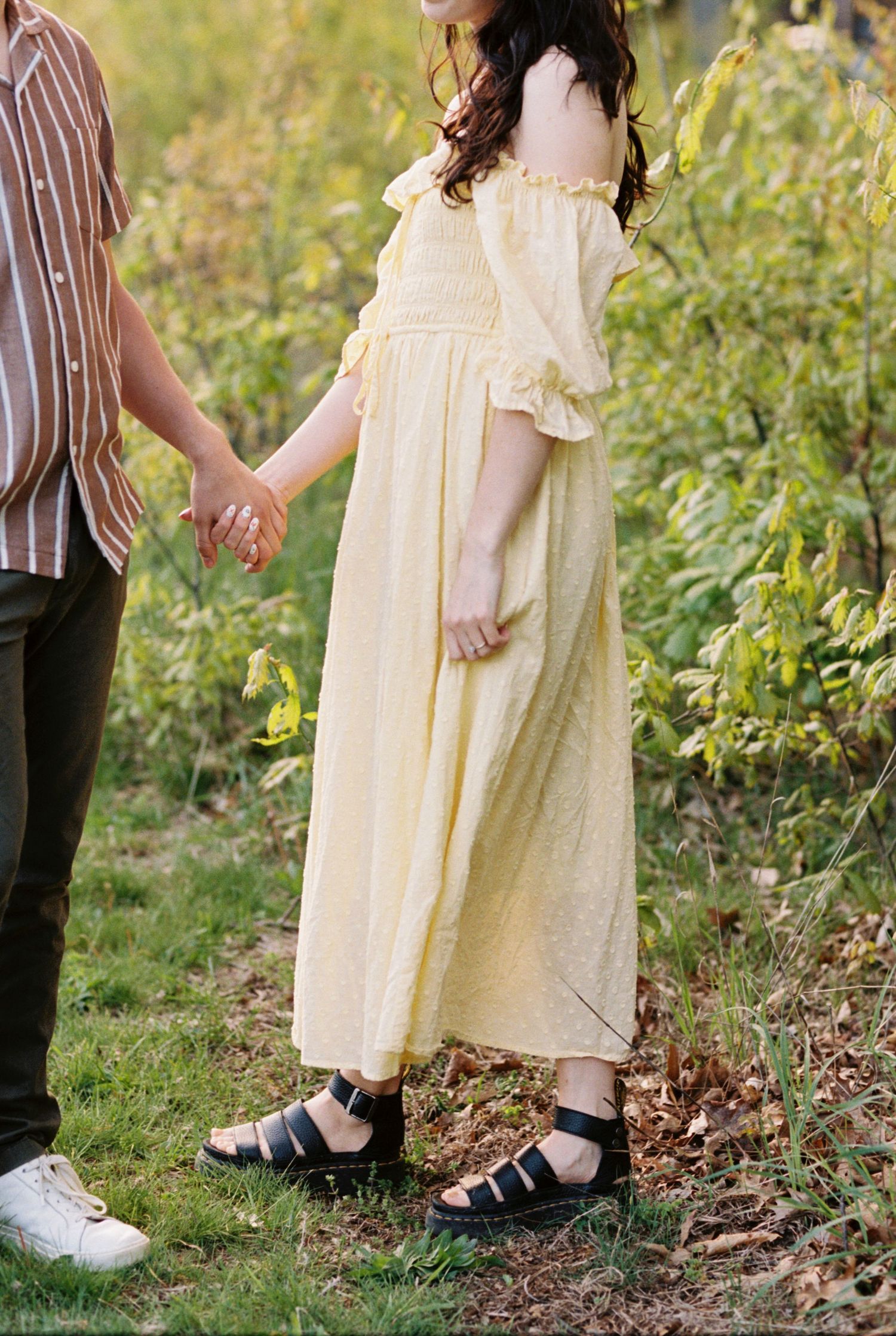 Person wearing a light yellow off-shoulder maxi dress with ruffled sleeves and black sandals holds hands in a garden setting.