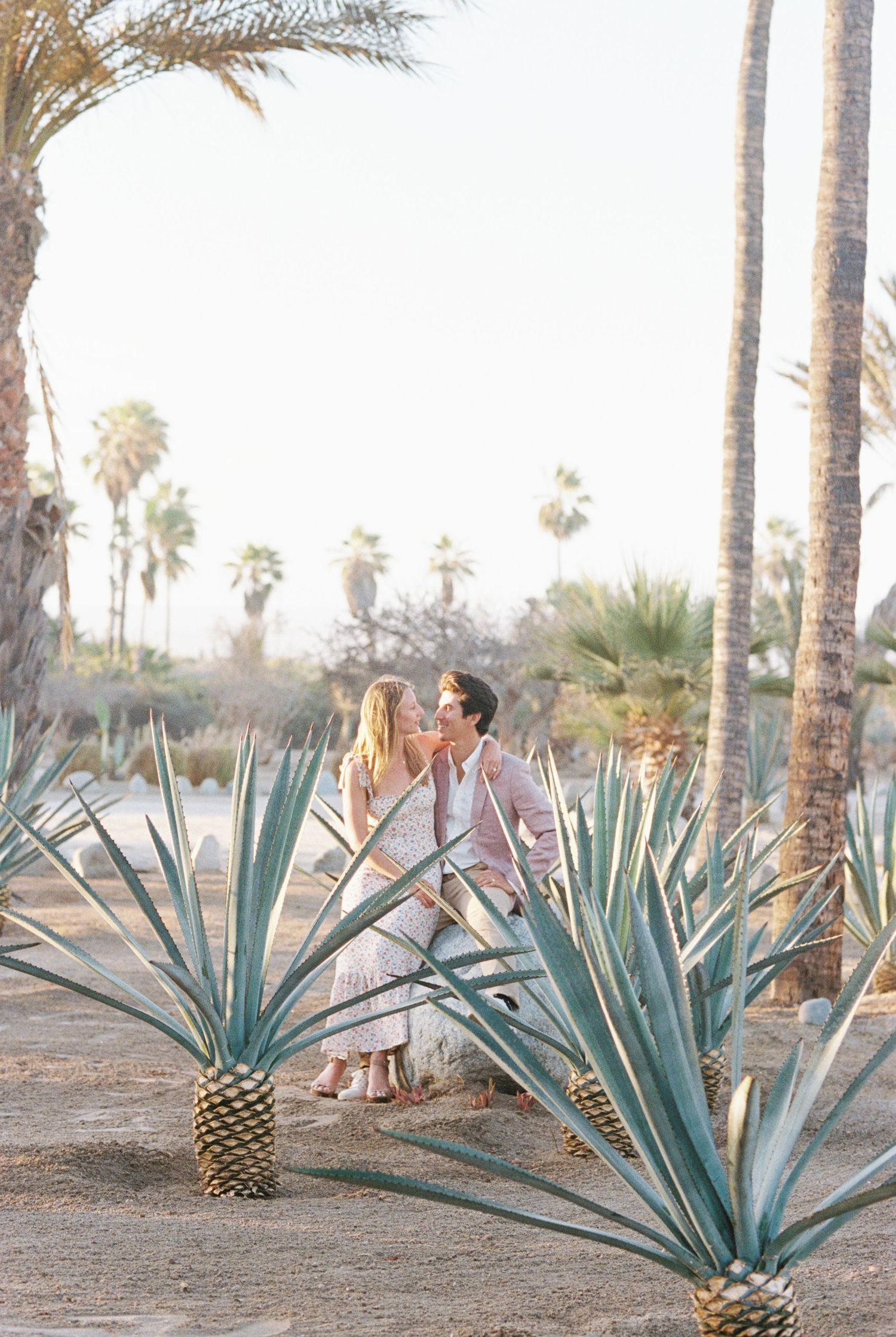 A couple in white outfits embracing among agave plants and palm trees in a desert garden at sunset. Kirstie Whitaker Photography