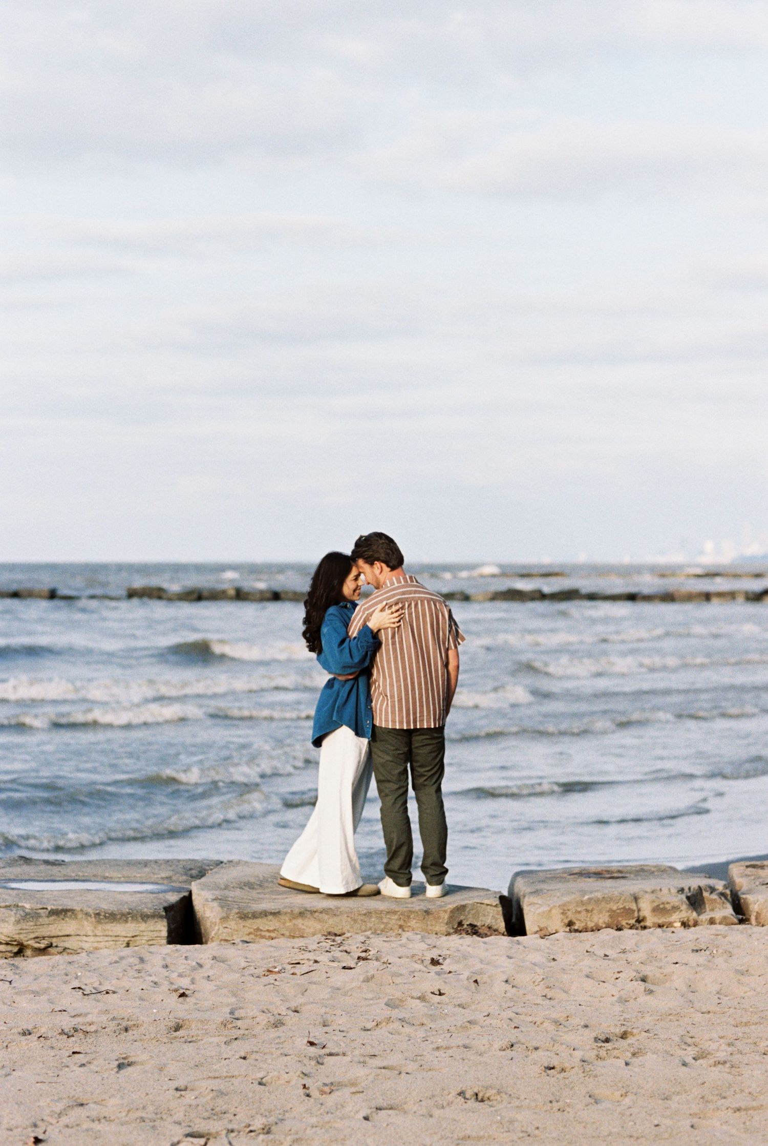 A romantic couple embraces while standing on a beach at sunset with gentle waves and rocks in the background.