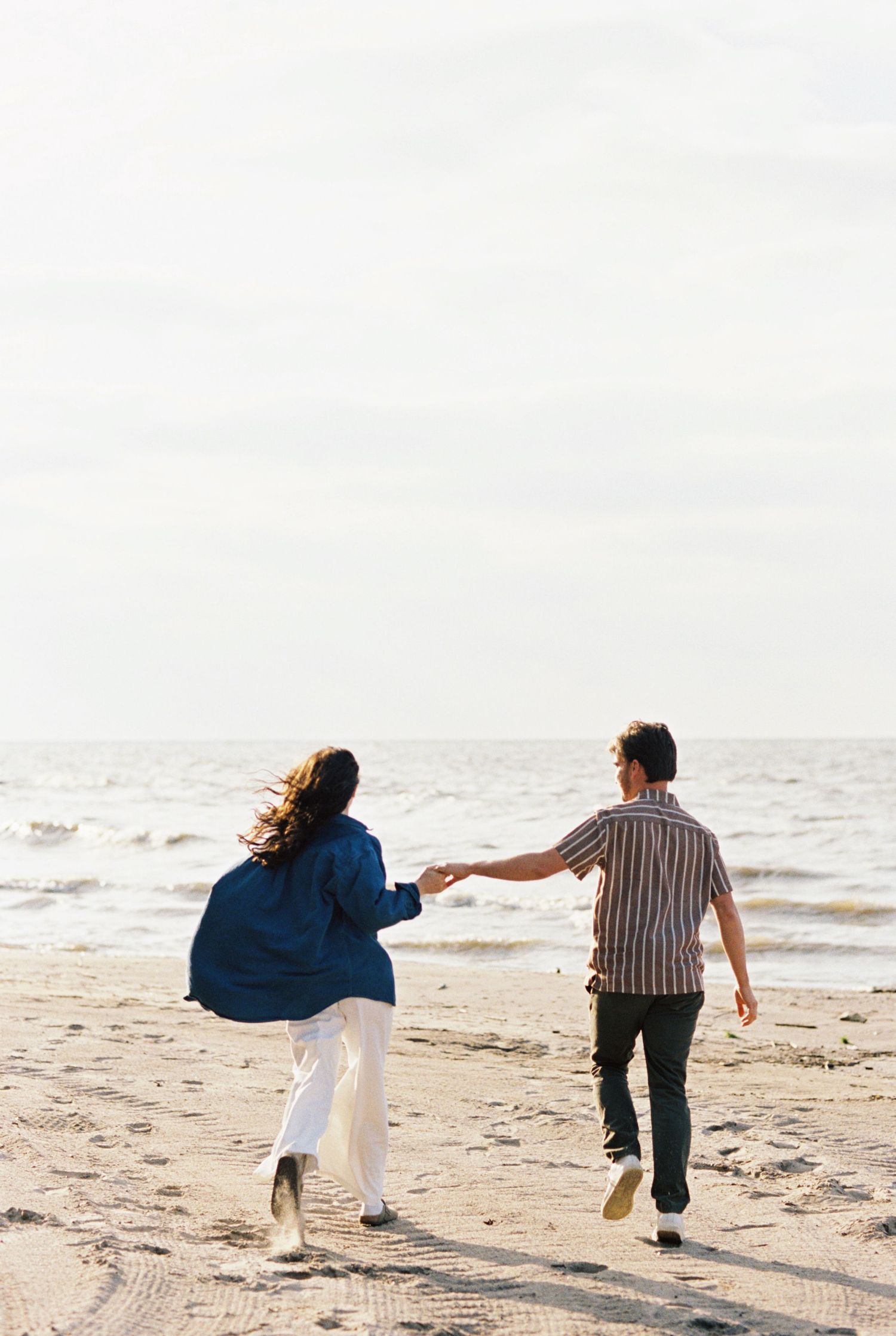 Two people holding hands while running together on a sandy beach by the ocean on an overcast day.