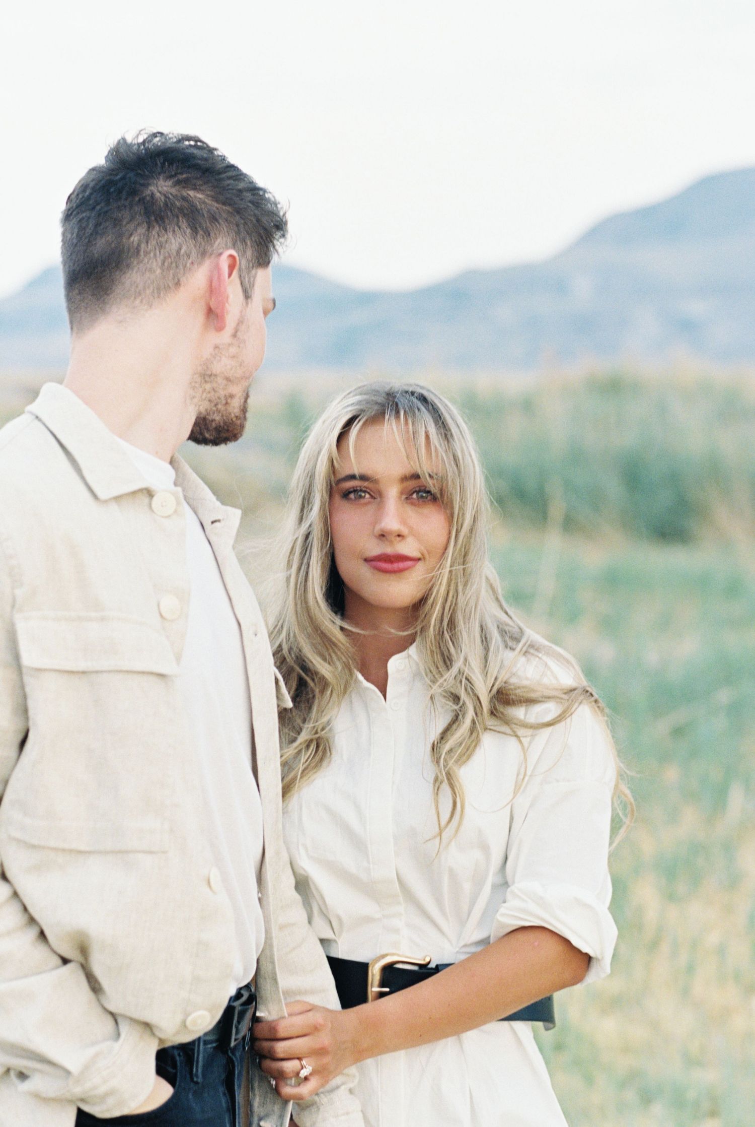 Couple in white outfits sharing an intimate moment outdoors with mountains and tall grass in the background.