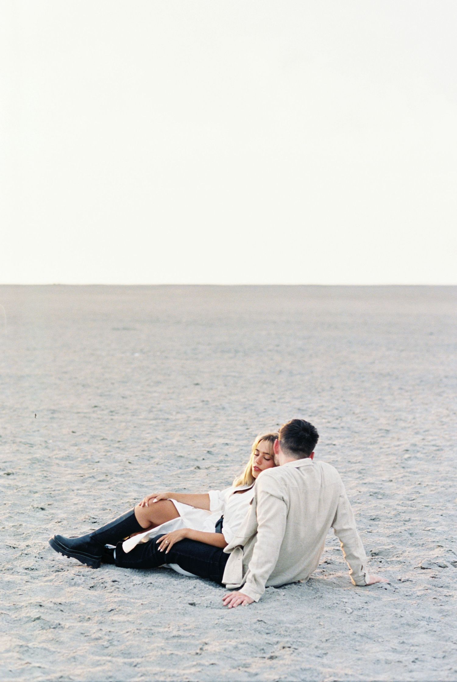 A couple sits intimately together on an empty beach with misty gray skies in soft, ethereal lighting.