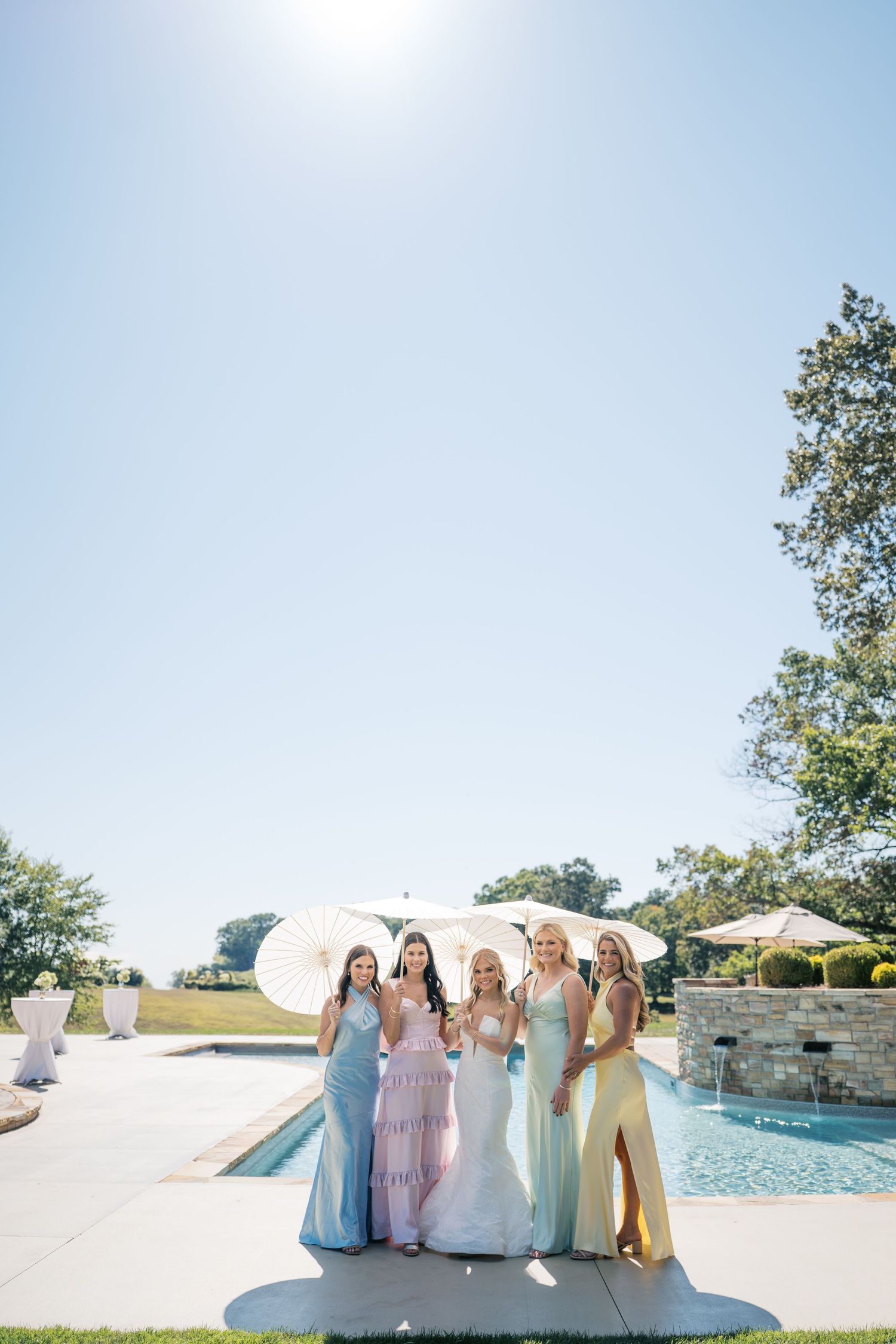 Bridesmaids in pastel colored dresses gather around a luxurious outdoor swimming pool on a sunny day.