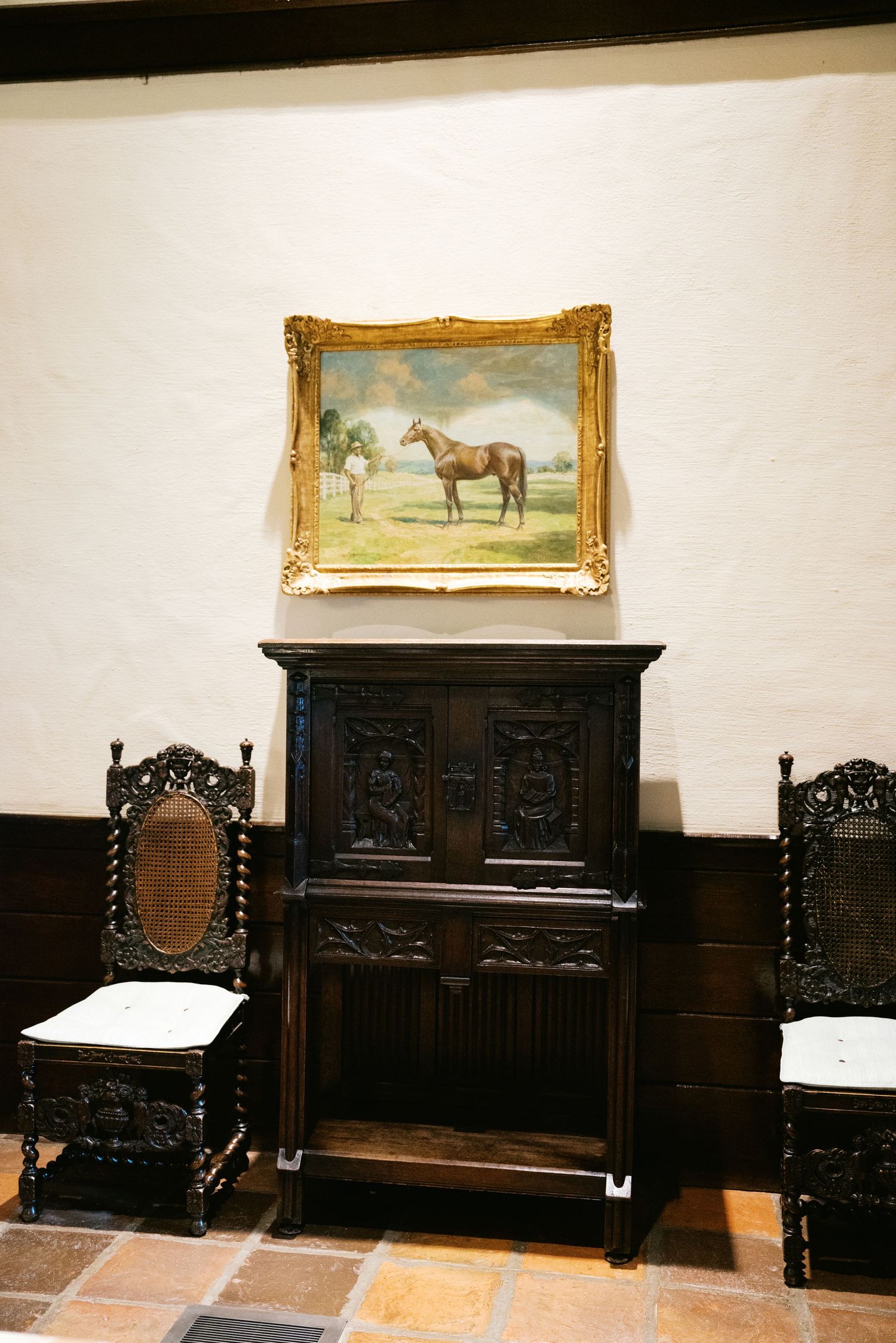 Antique dark wood furniture with ornate carved chairs and cabinet beneath a framed horse painting in gold frame.