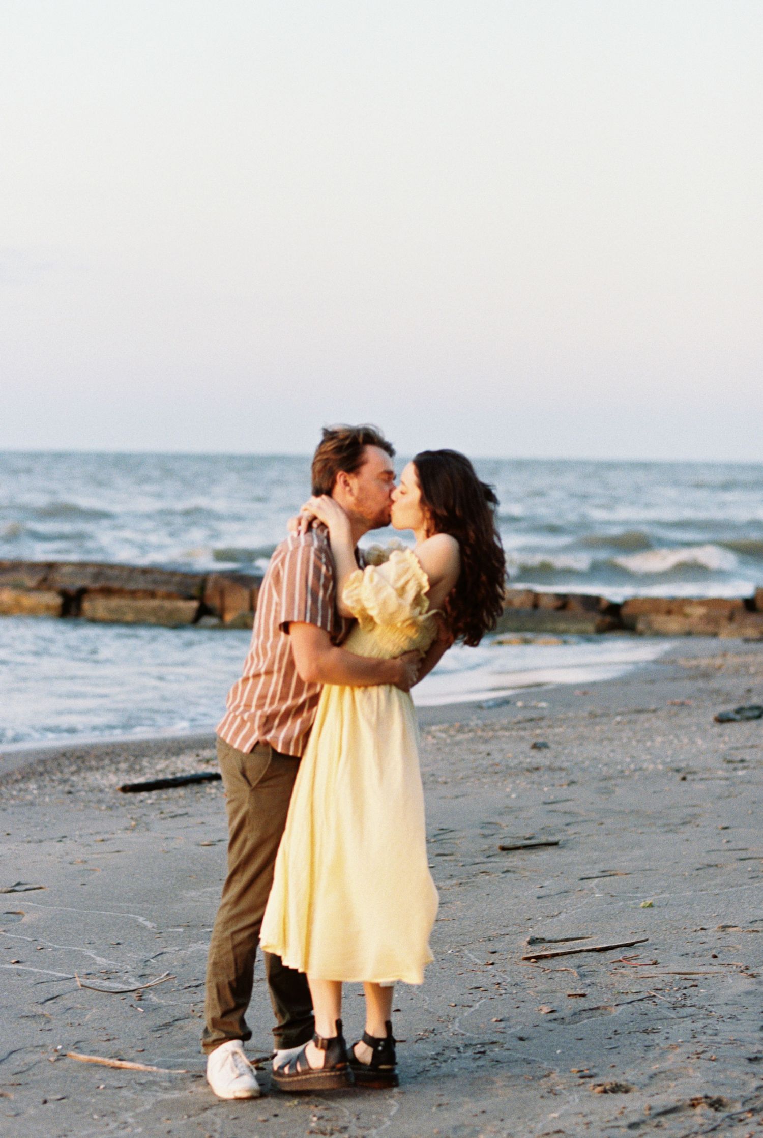A romantic couple embracing on a scenic beach with rocky outcrops and ocean waves at dusk.