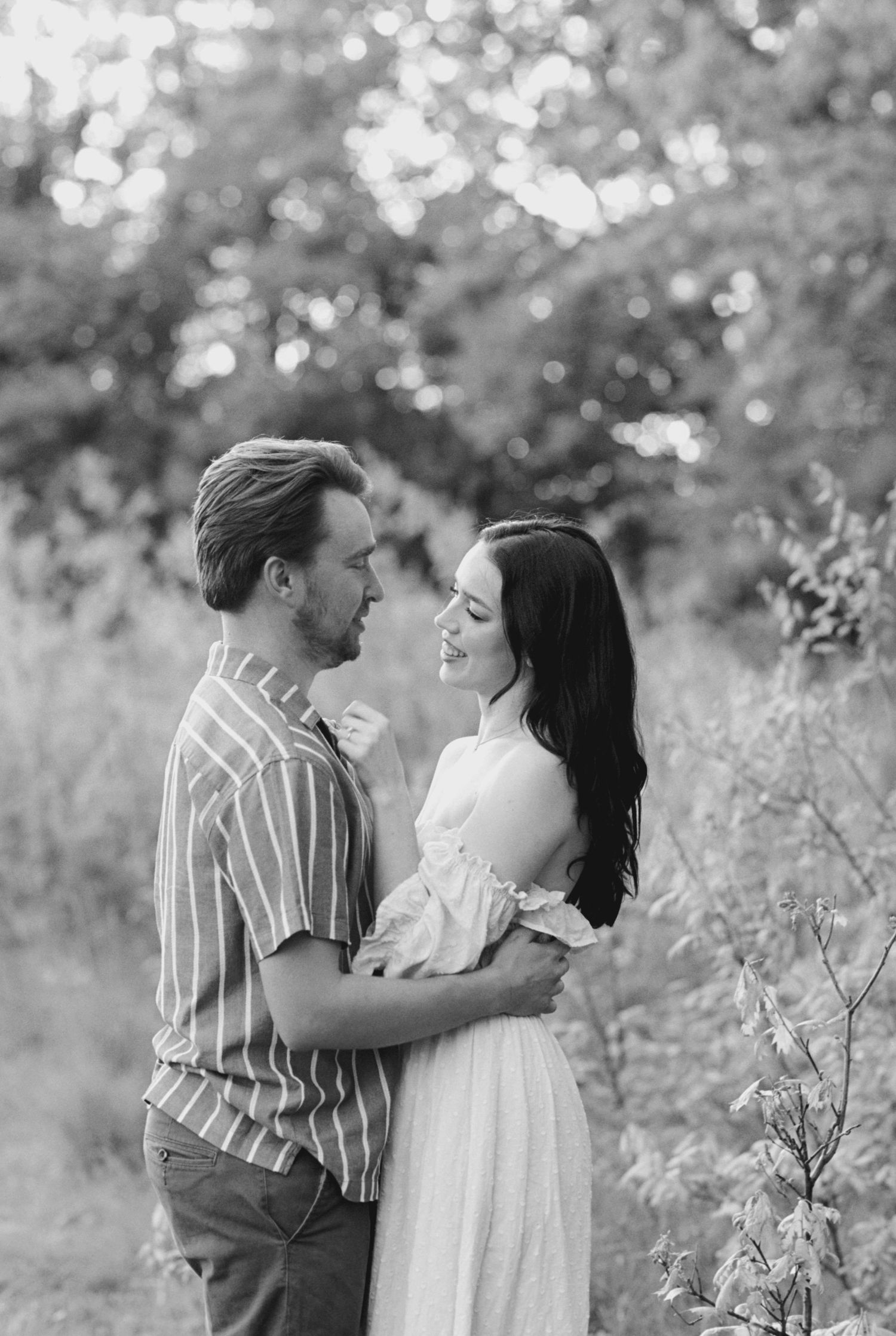 A romantic black and white portrait of a couple embracing in a sunlit meadow with bokeh effect in the background.