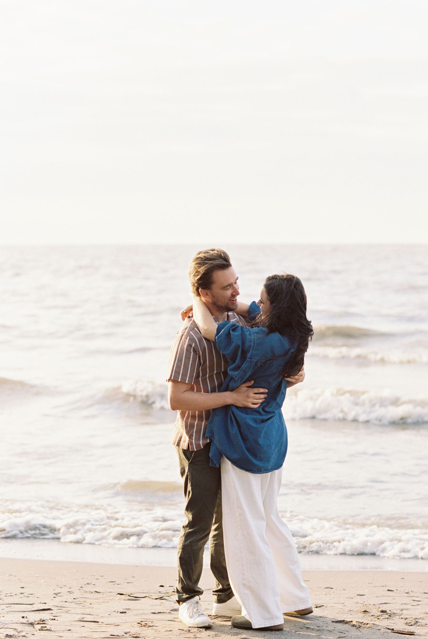 Romantic couple embracing on sandy beach at sunset with gentle waves rolling in behind them.