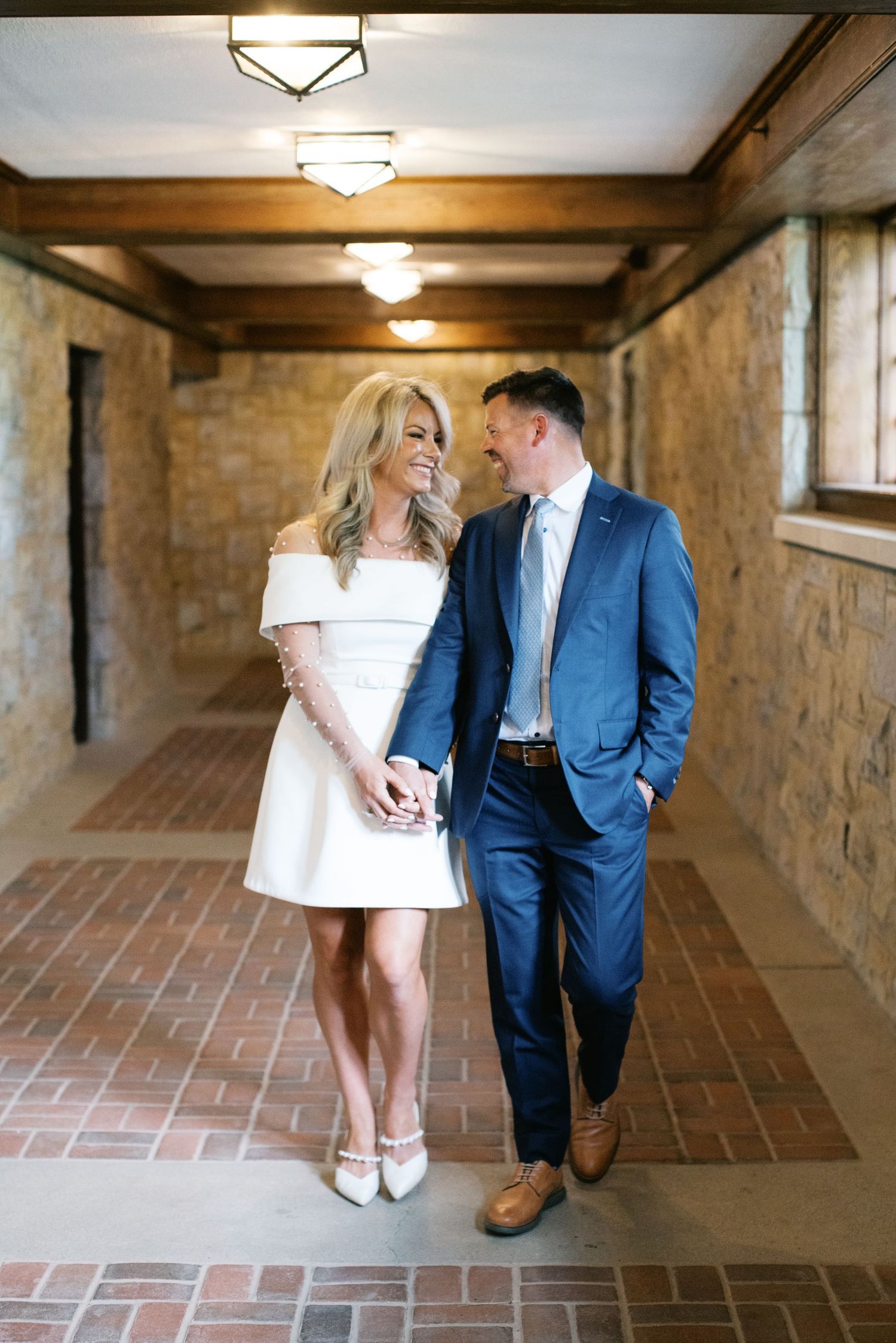 A couple shares an intimate moment in a stone hallway, with her in white dress and him in blue suit.