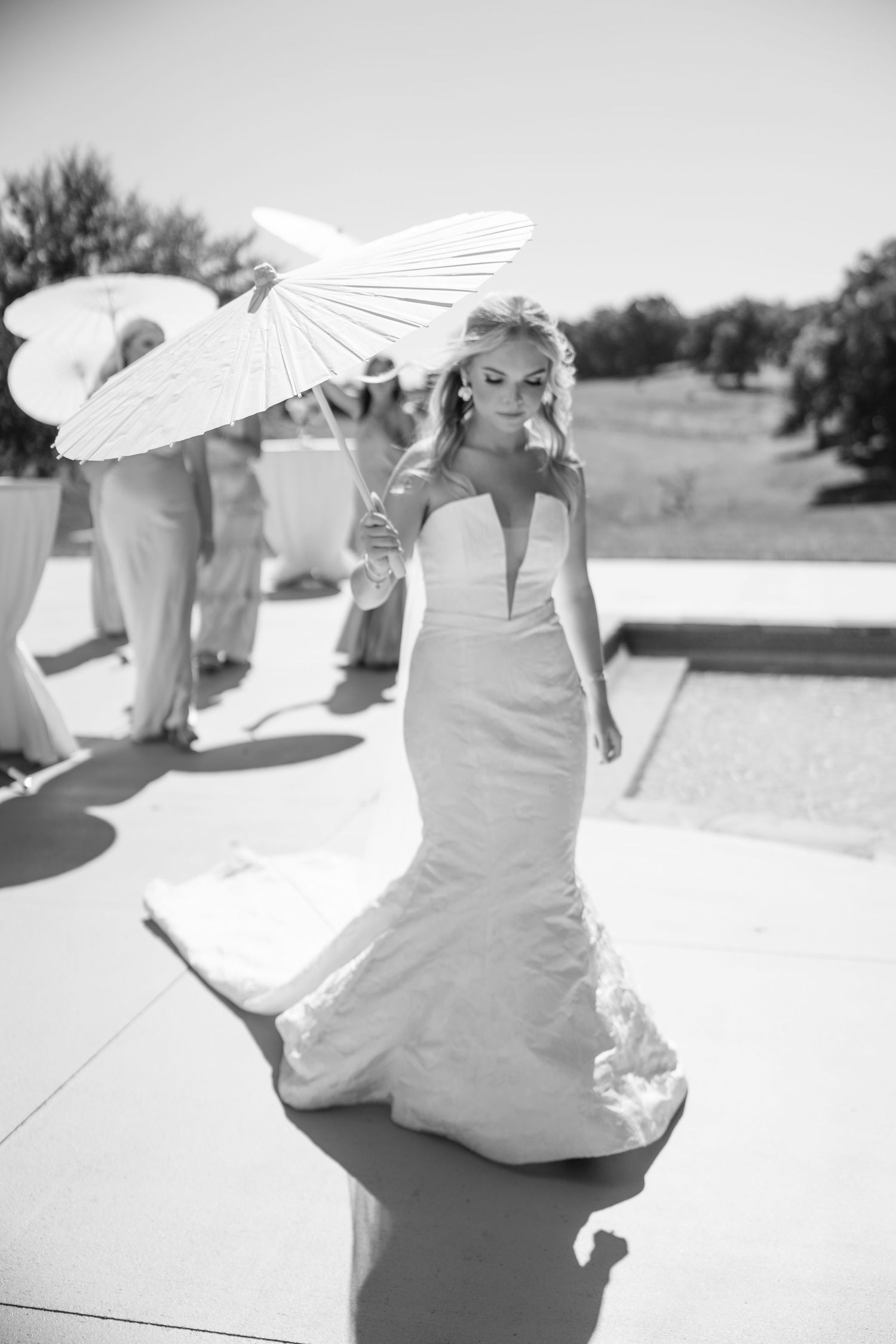 A bride in a fitted mermaid wedding gown holds a white parasol while posing outdoors in black and white photography.