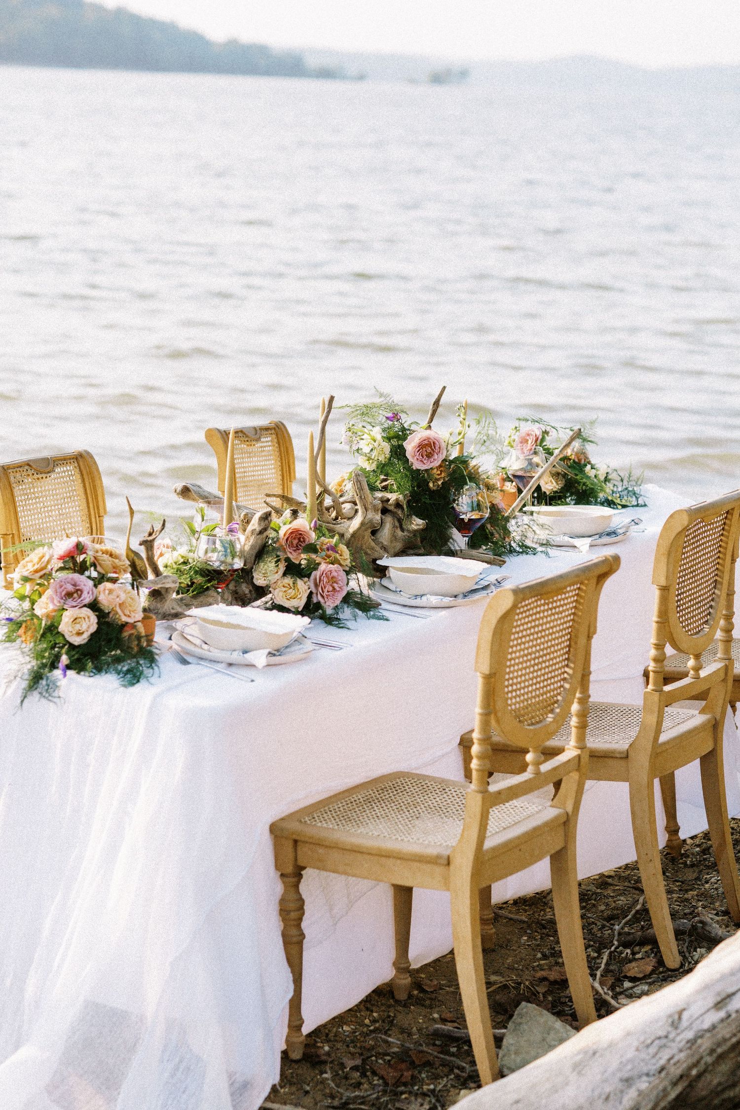 Elegant beach dining setup with white tablecloth, cane chairs, and romantic floral centerpieces overlooking the water.