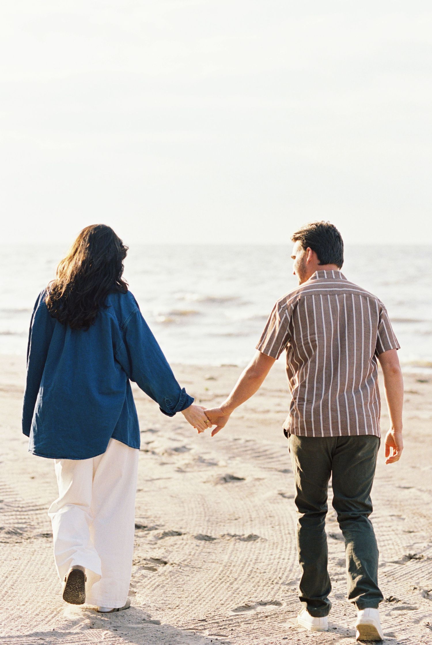 A couple holds hands while walking together along a serene beach at sunset.