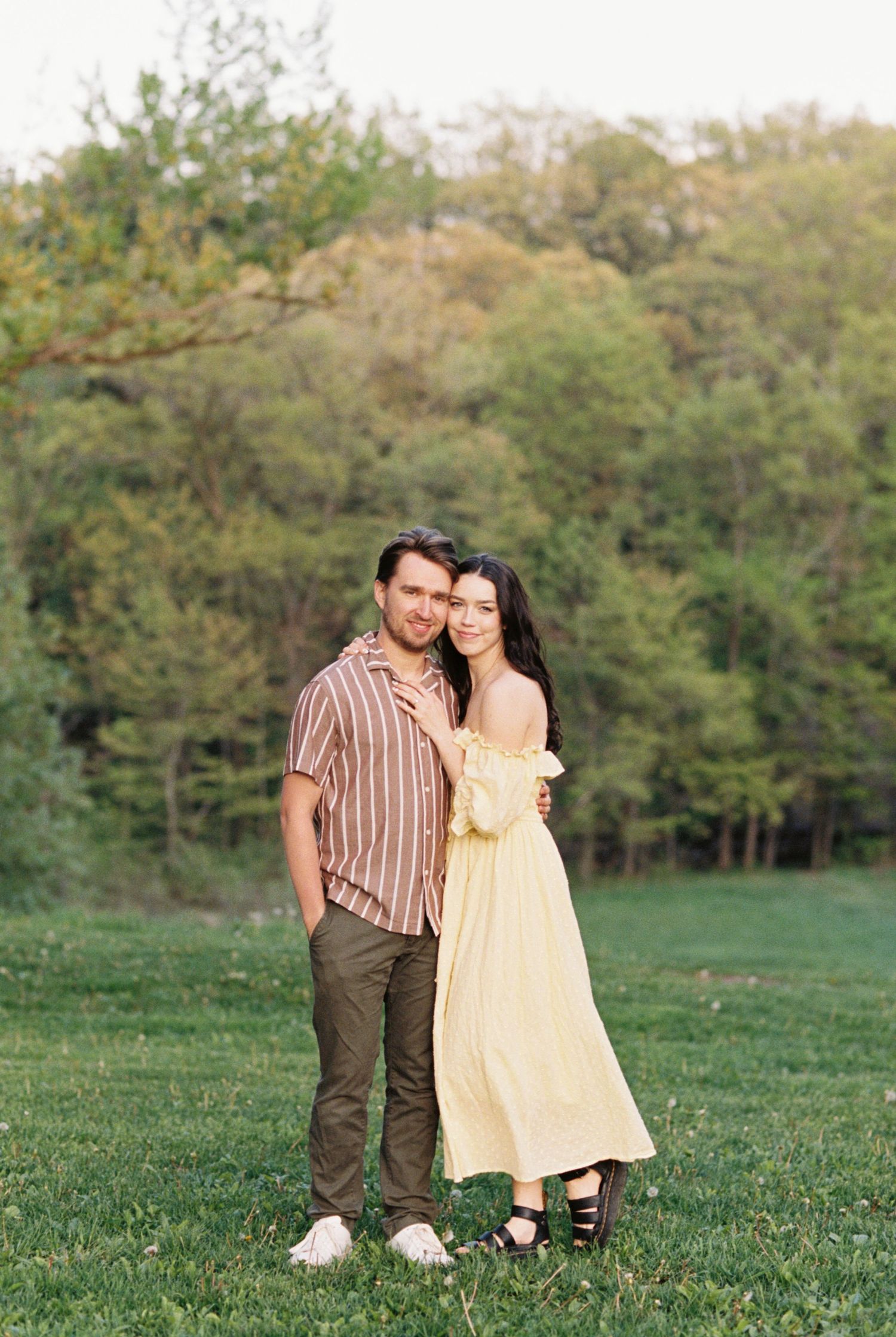 A couple stands together on a grassy field with trees in the background during golden hour.
