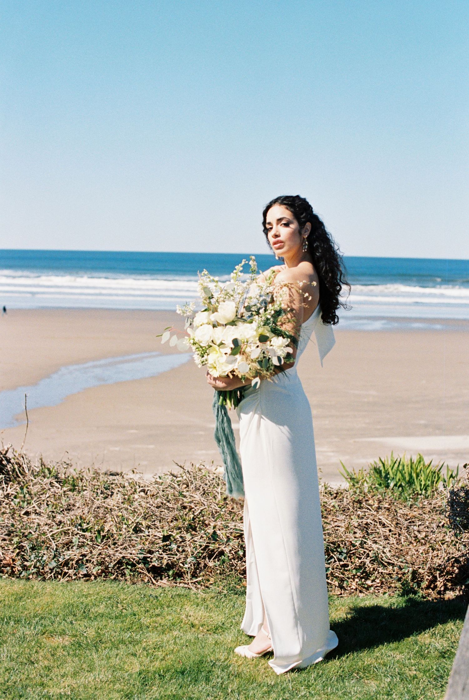 Elegant bride in white dress holds lush bouquet of white flowers while standing on grassy overlook at sunny beach.