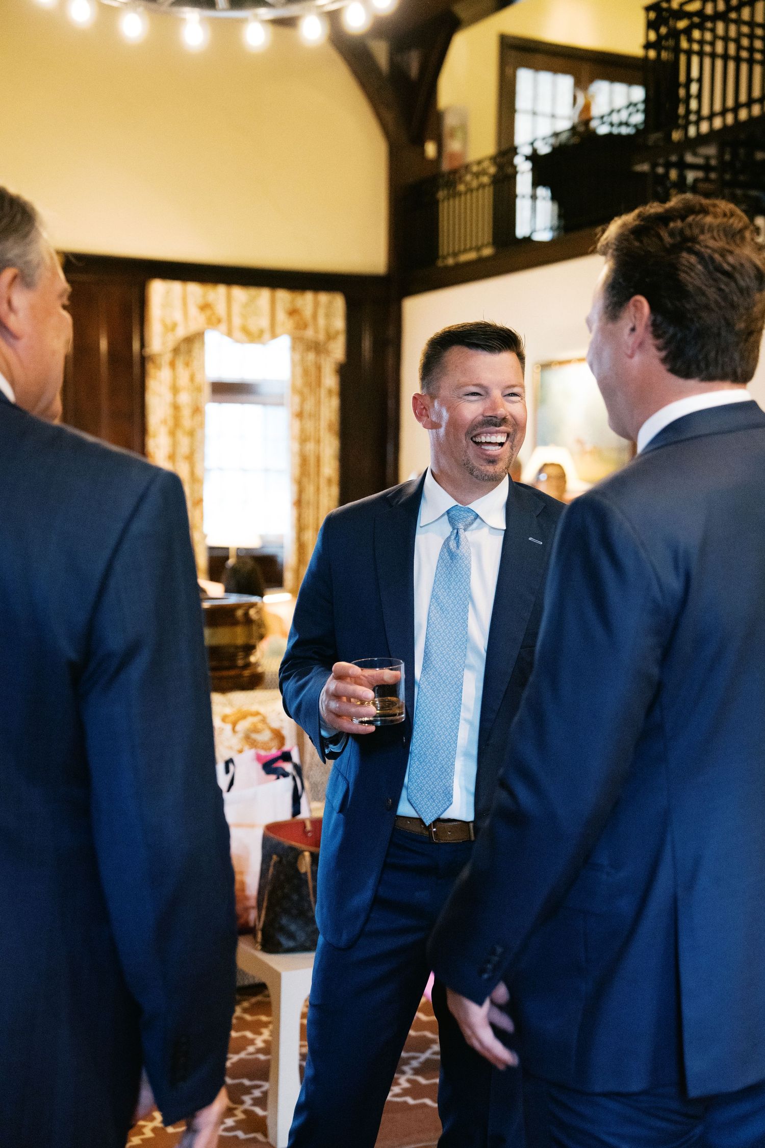 Three men in navy suits and light blue ties share a conversation at an elegant indoor venue with warm lighting.