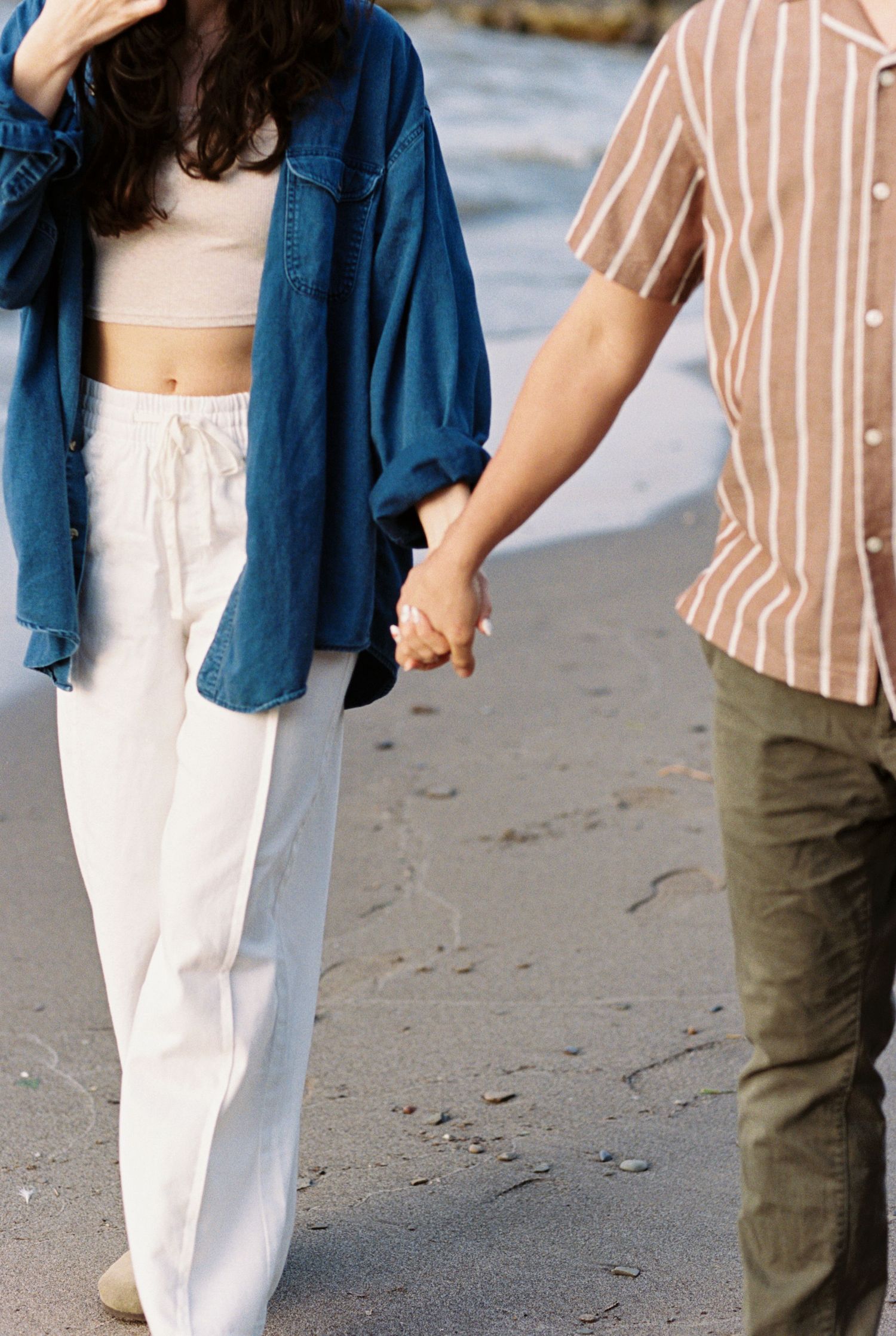 Couple holding hands while walking on beach wearing casual summer outfits of denim, stripes and white pants.