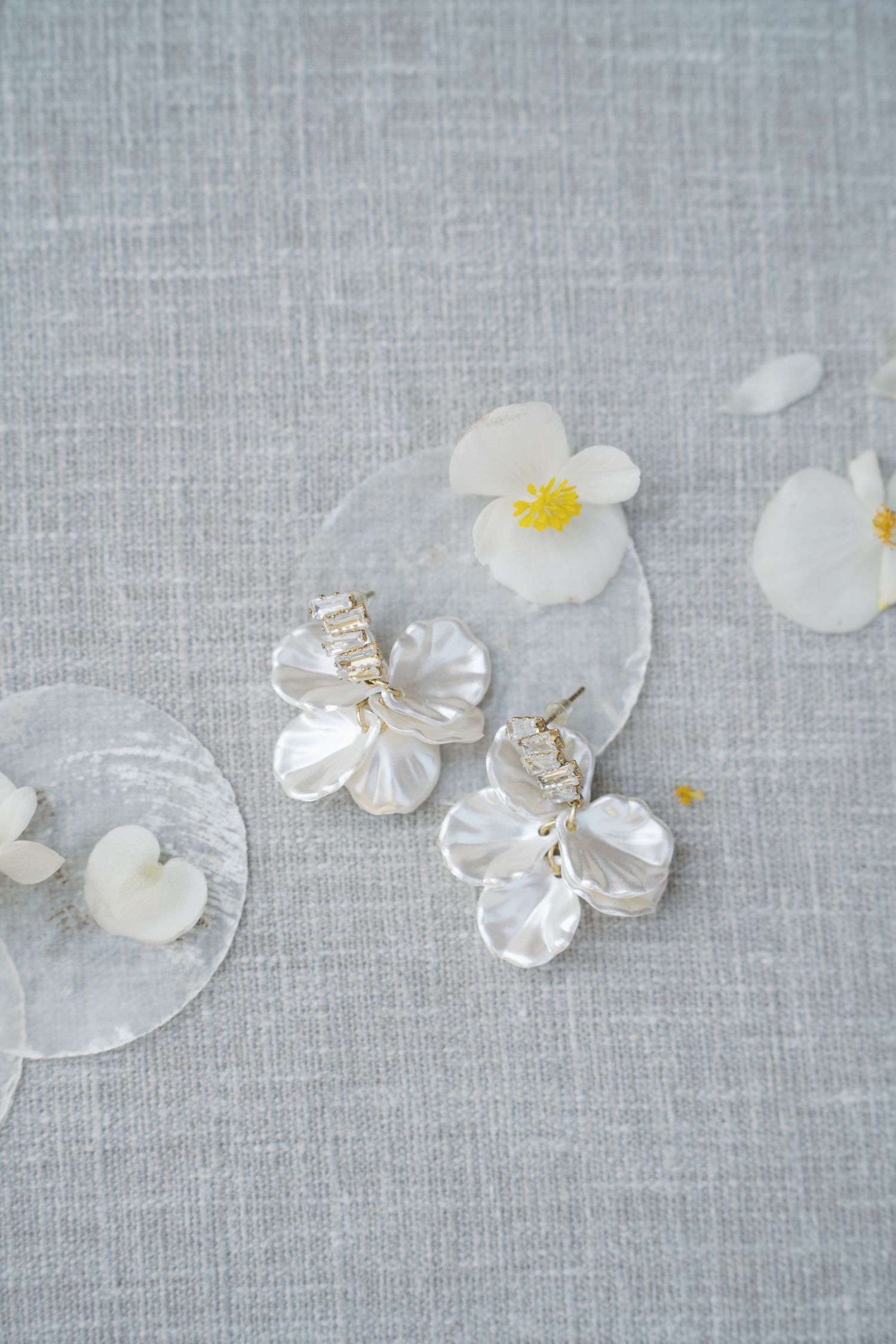White begonia flower petals and yellow stamens scattered artistically on light gray linen textile background.