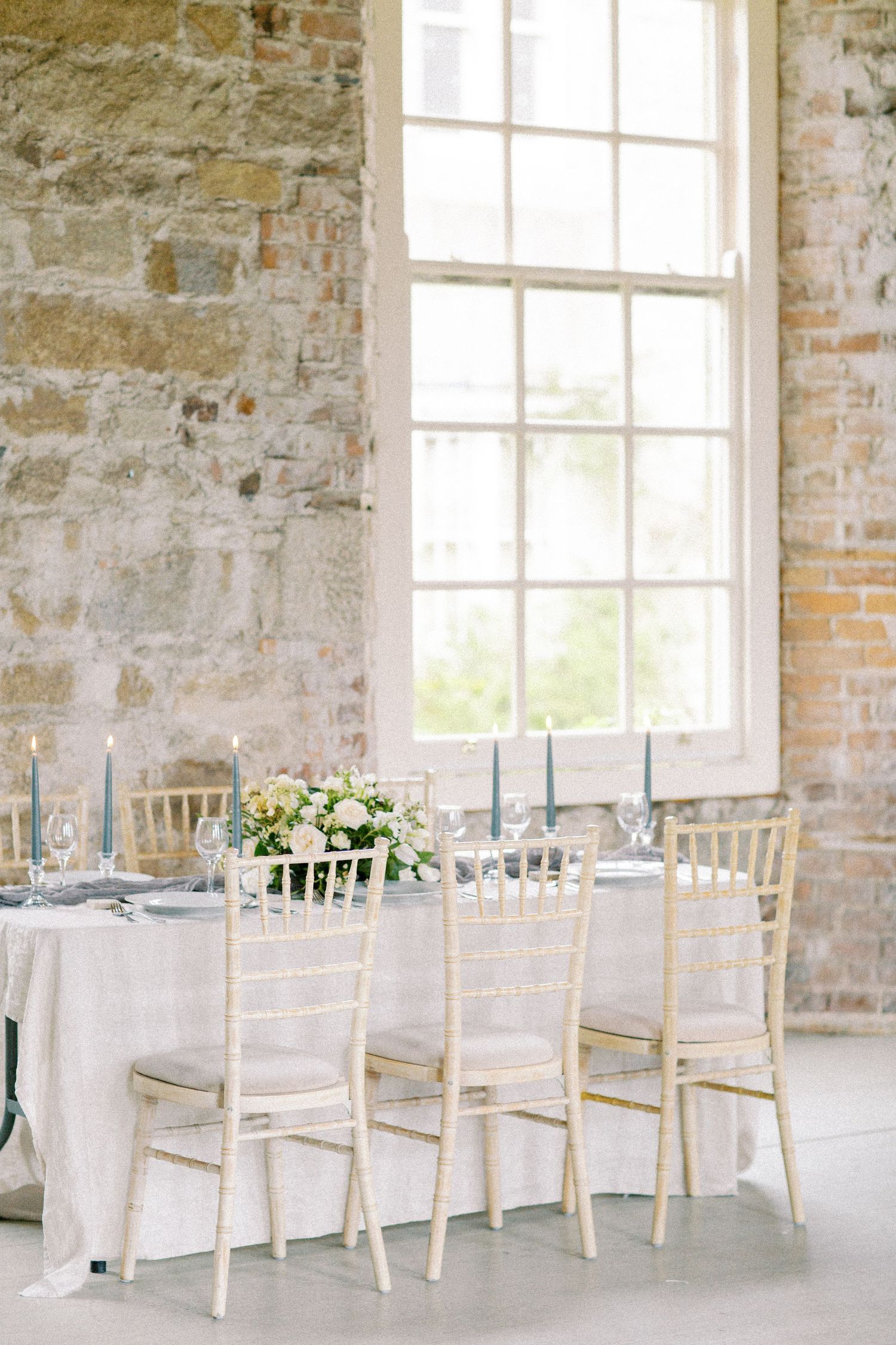 An elegant wedding reception table setting with white chiavari chairs and blue accents in a rustic stone room. Kirstie Whitaker Photography