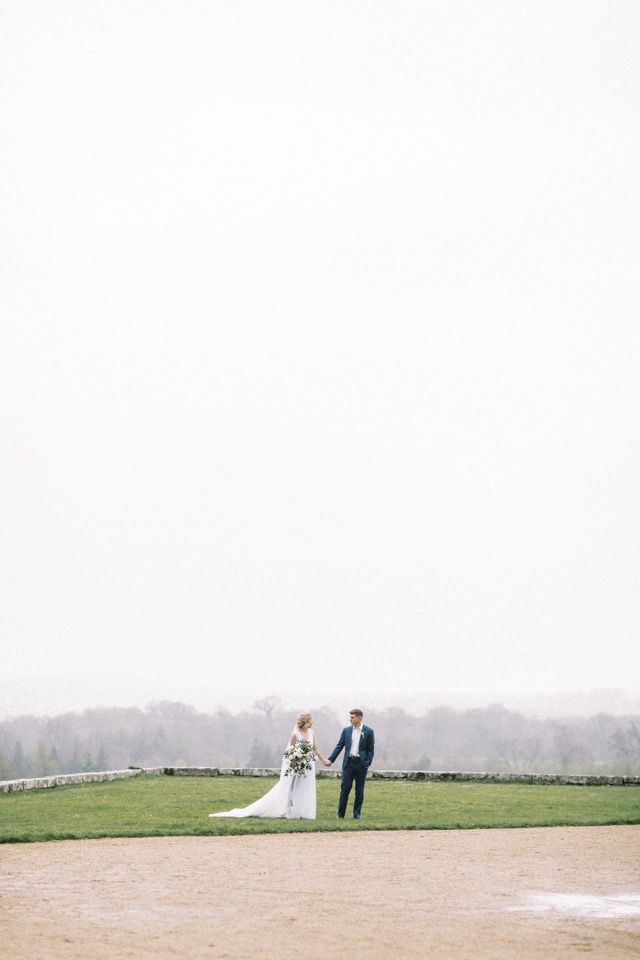 A couple in wedding attire stands on a grassy hilltop overlooking a misty countryside landscape in the distance.