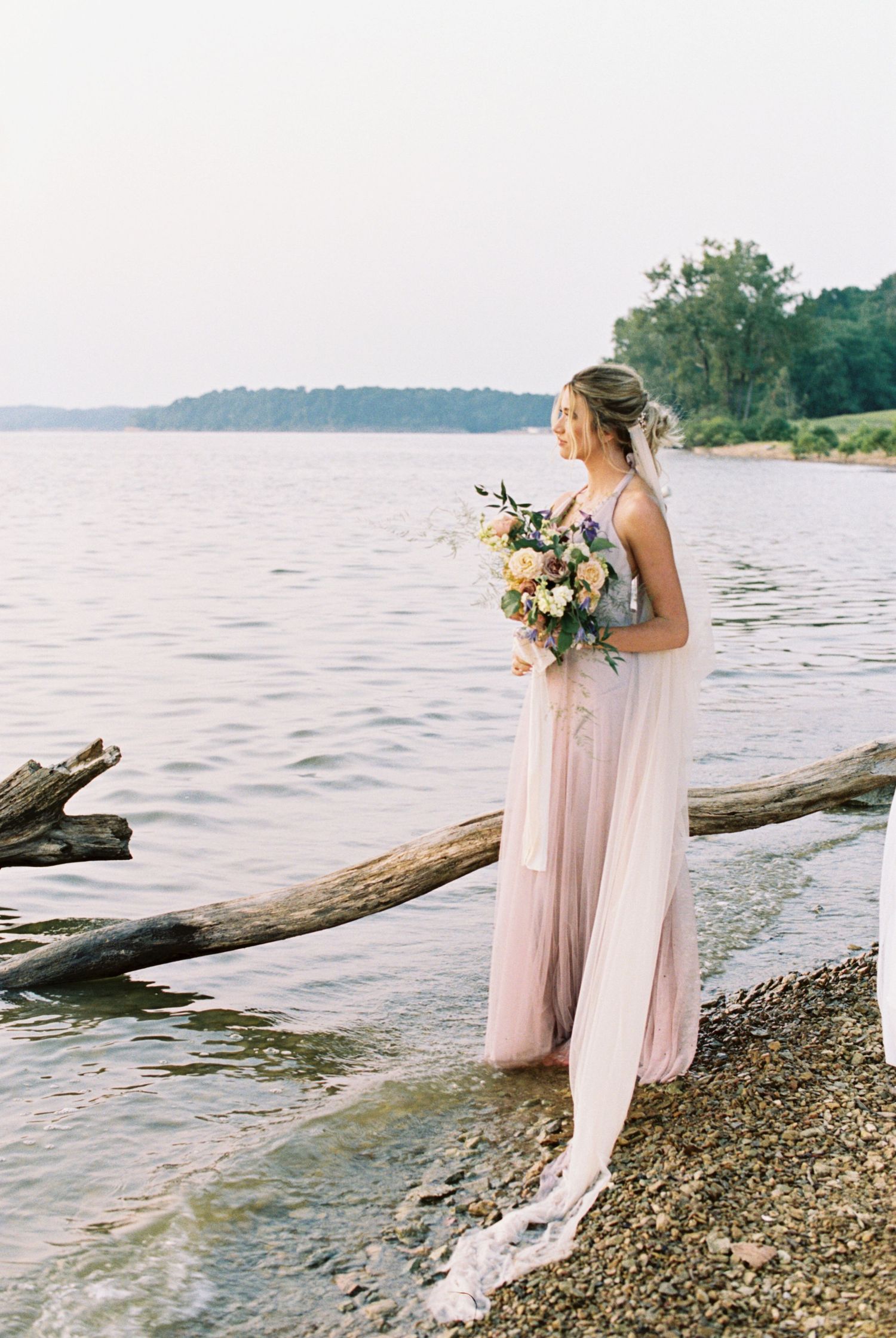 A person in a flowing blush dress holds a floral bouquet while standing on a rocky lakeshore during sunset.
