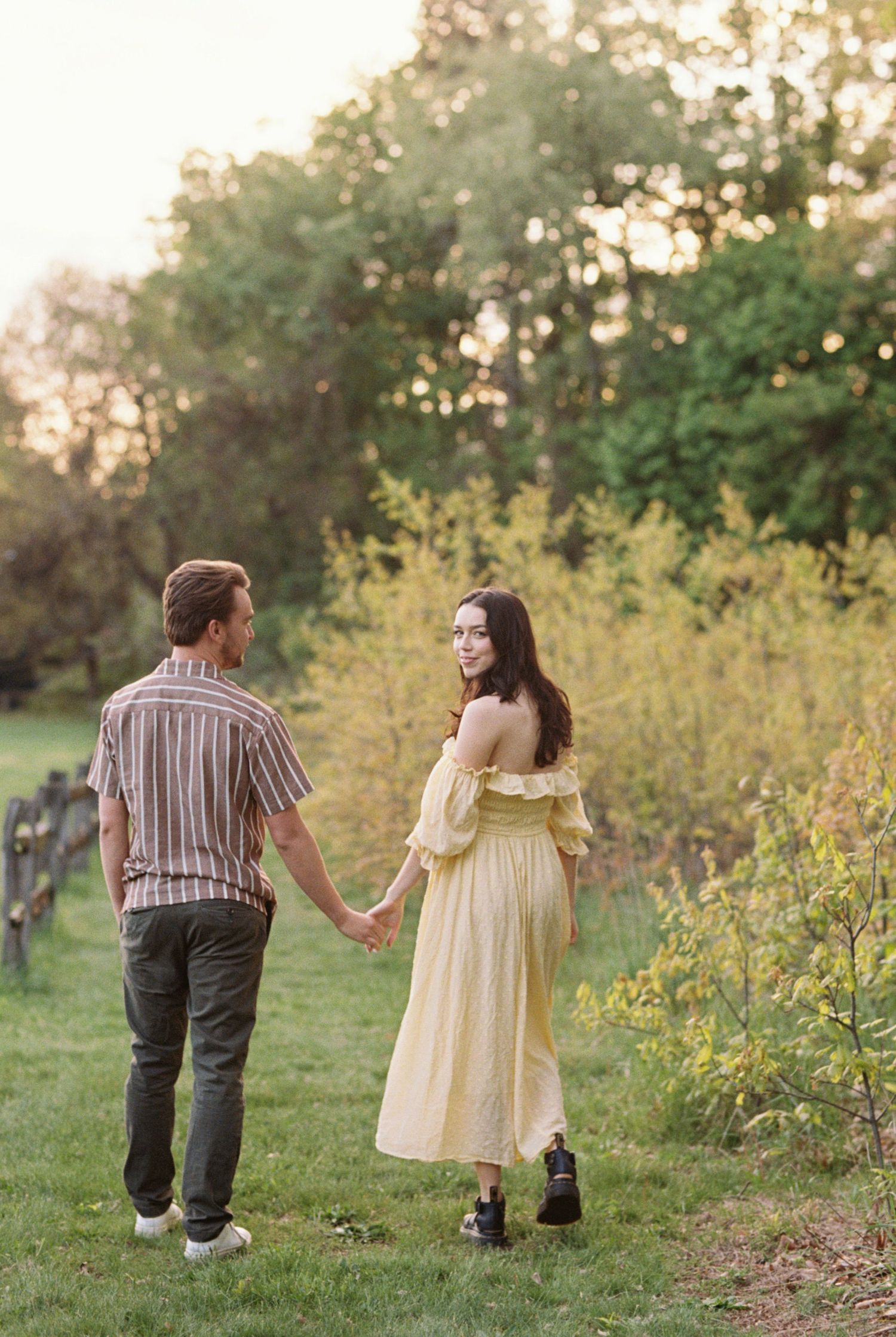 A couple holds hands while walking through a meadow at sunset, her in a yellow vintage dress and him in stripes.