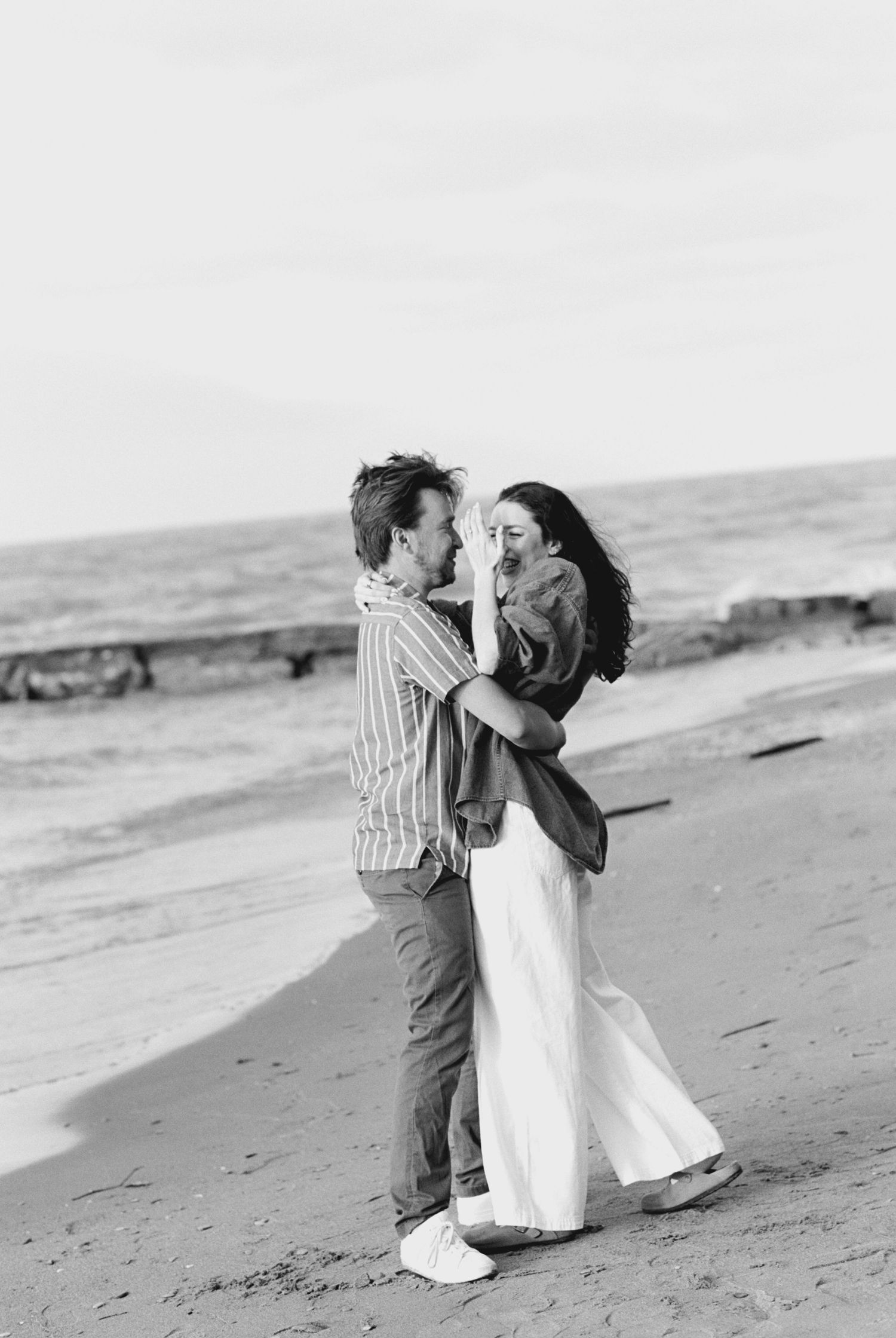 A romantic black and white beach portrait showing a couple embracing and laughing together on the sand by the ocean.