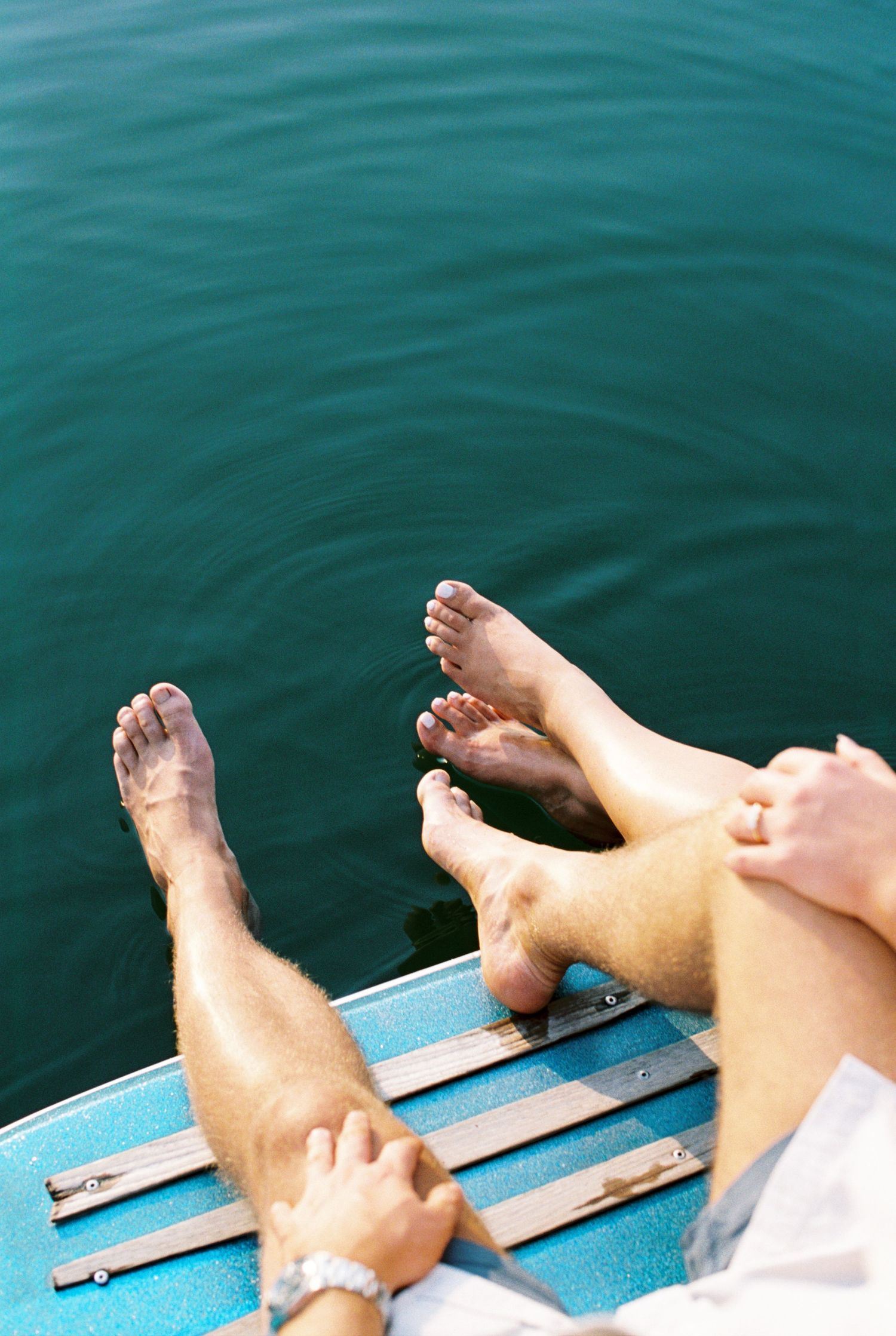 Bare feet dangling over turquoise water from the edge of a weathered blue wooden boat on a calm summer day.
