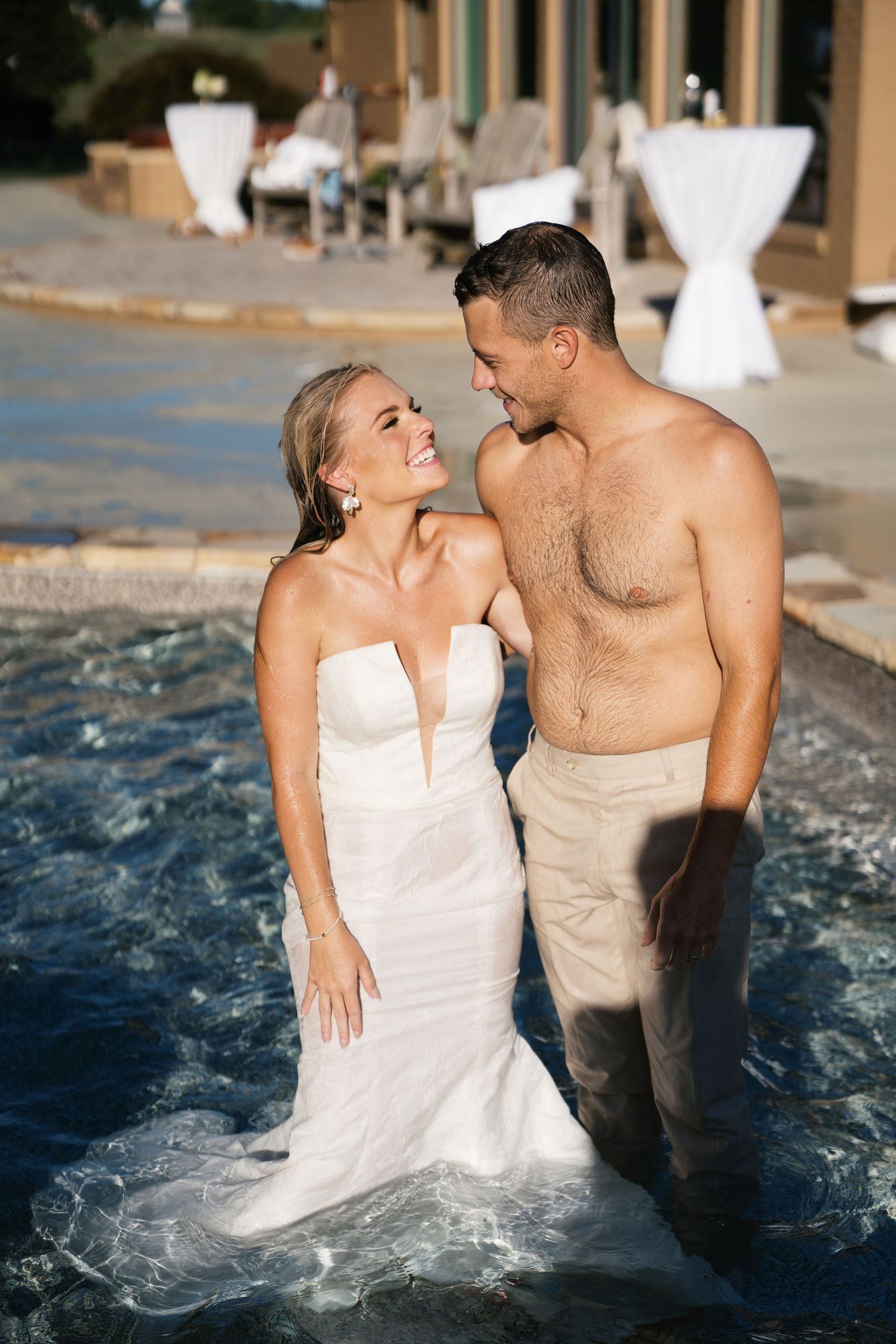 A couple stands together in a pool during a wedding celebration with cocktail tables visible in the background.