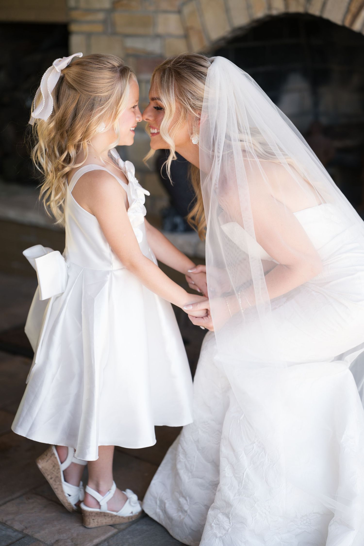A bride in white wedding dress and veil shares a tender moment with a small child in a white dress.