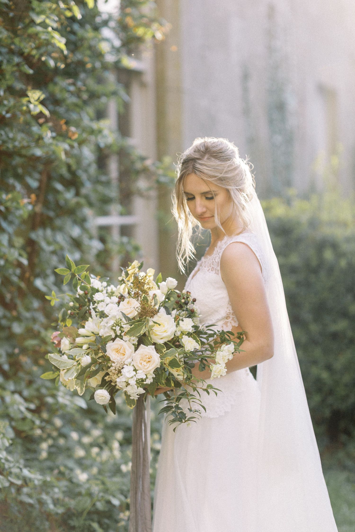 A bride in a white sleeveless gown holds a lush bouquet of white and blush roses against a garden backdrop.