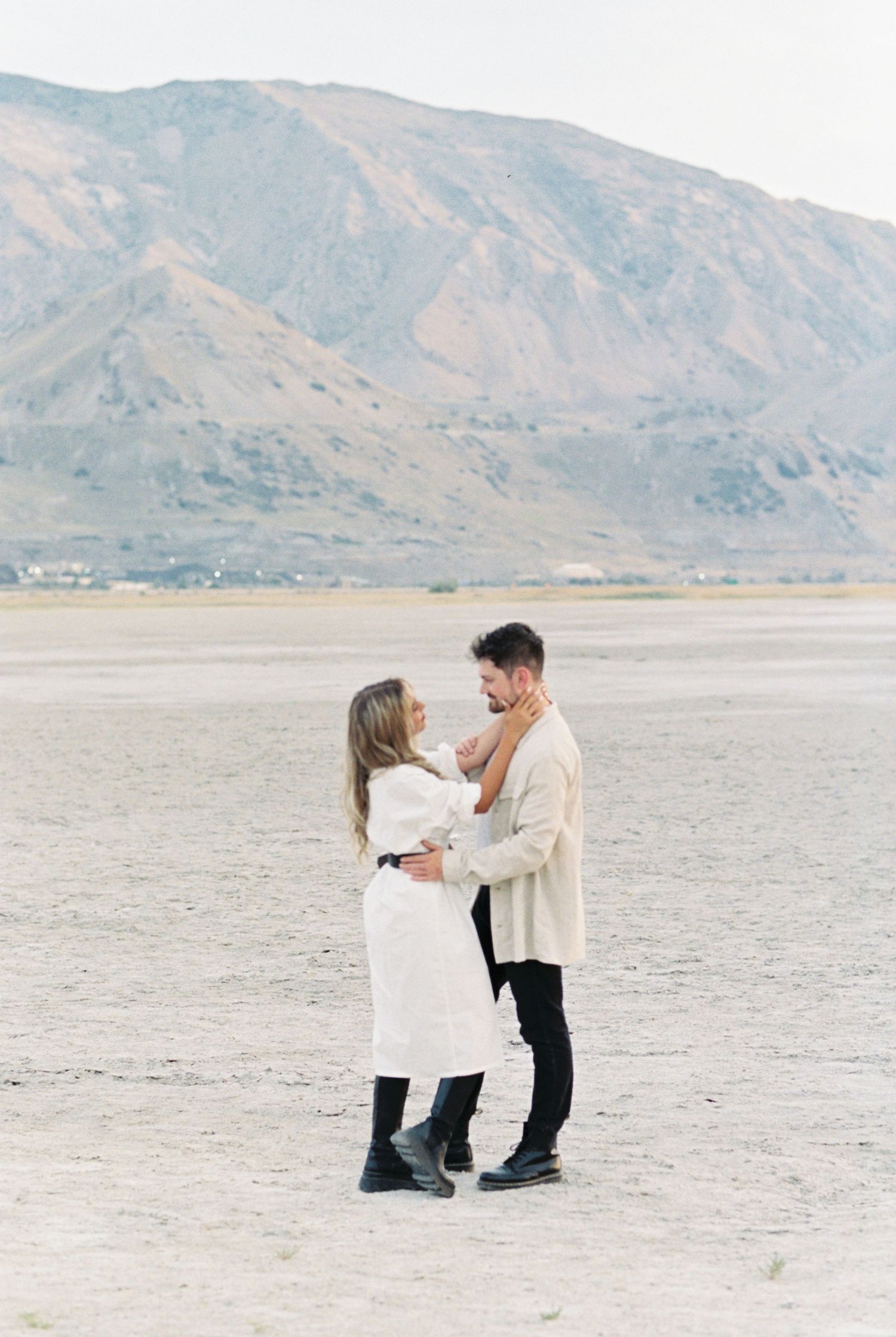 A couple embraces on a desert plain with mountainous terrain in the background during a soft, hazy day.