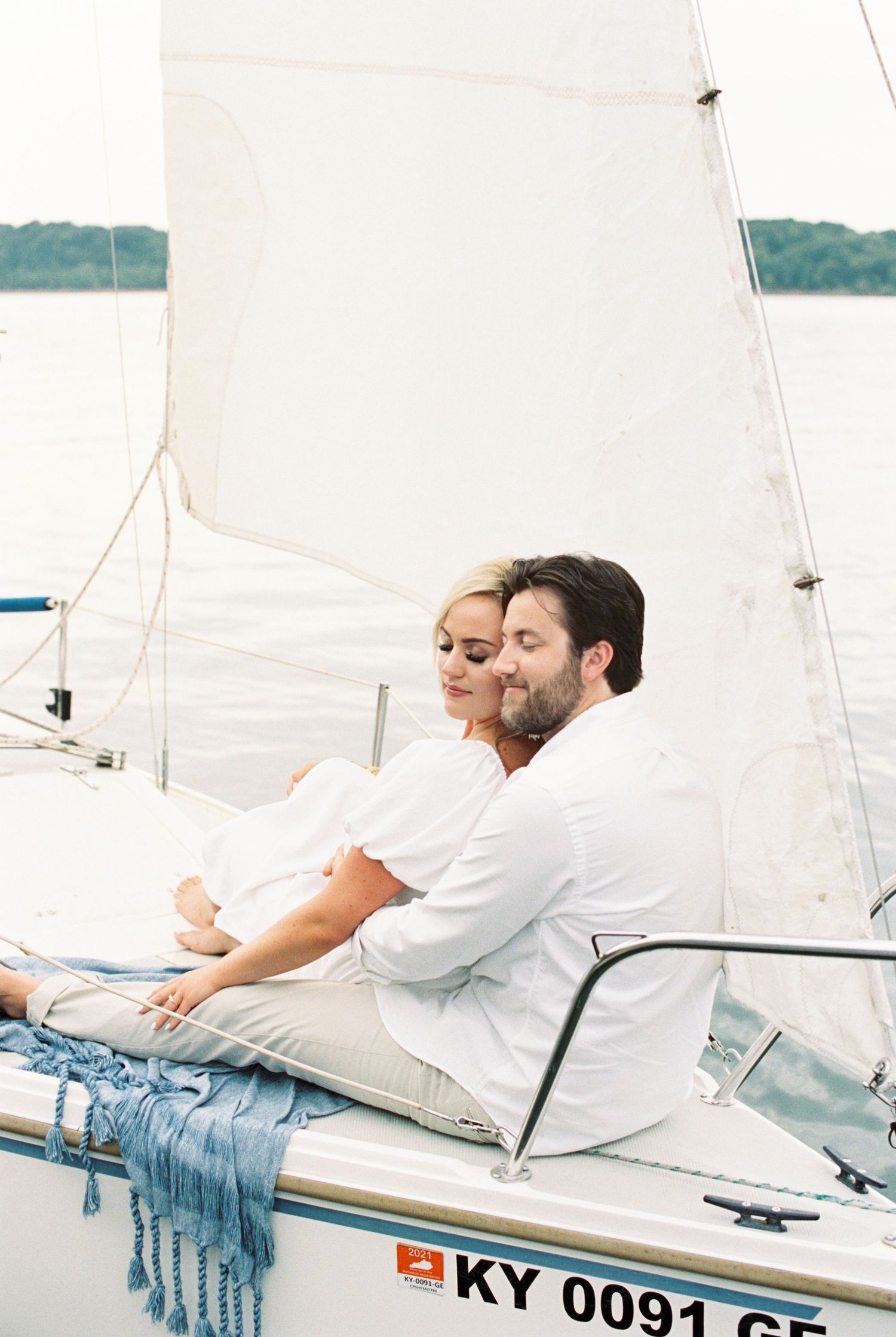 A couple relaxes together on a sailboat with white sails against a peaceful waterfront background.