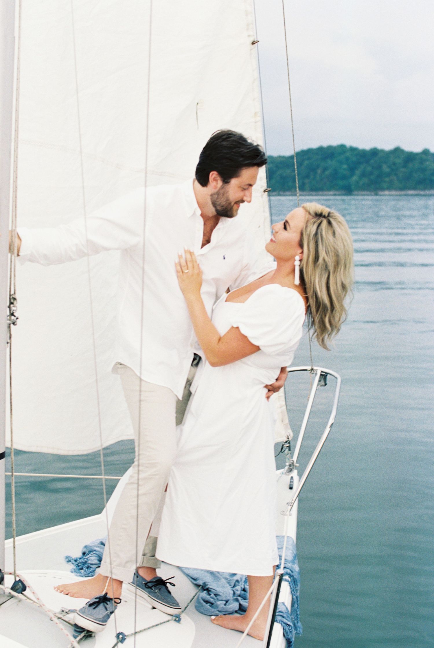 A romantic moment between a couple in white clothing on a sailboat deck against a serene water backdrop. Kirstie Whitaker Photography