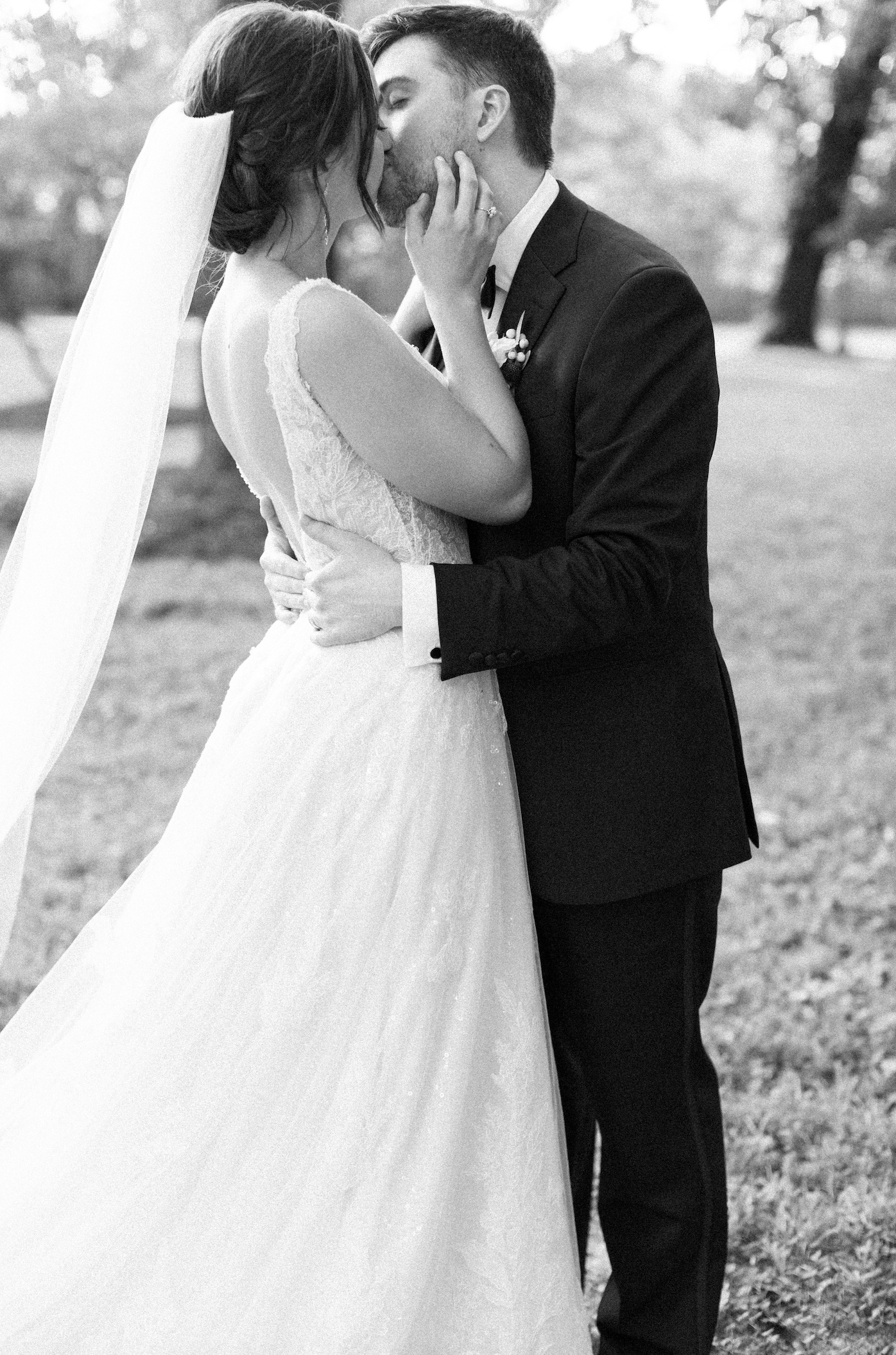 Black and white photo of a romantic wedding kiss between a bride in lace dress and groom in classic suit outdoors.