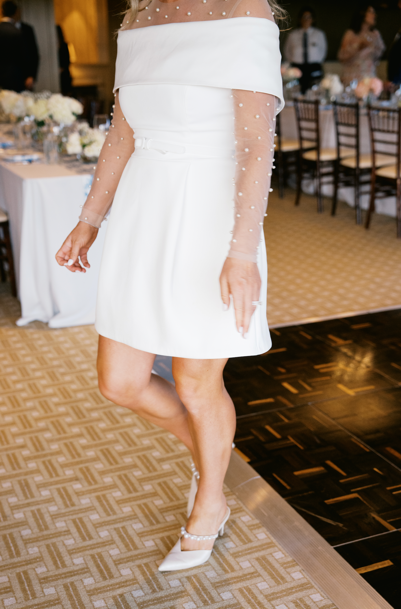A person in a short white off-shoulder dress and white heels stands on a patterned ballroom floor at a wedding reception.