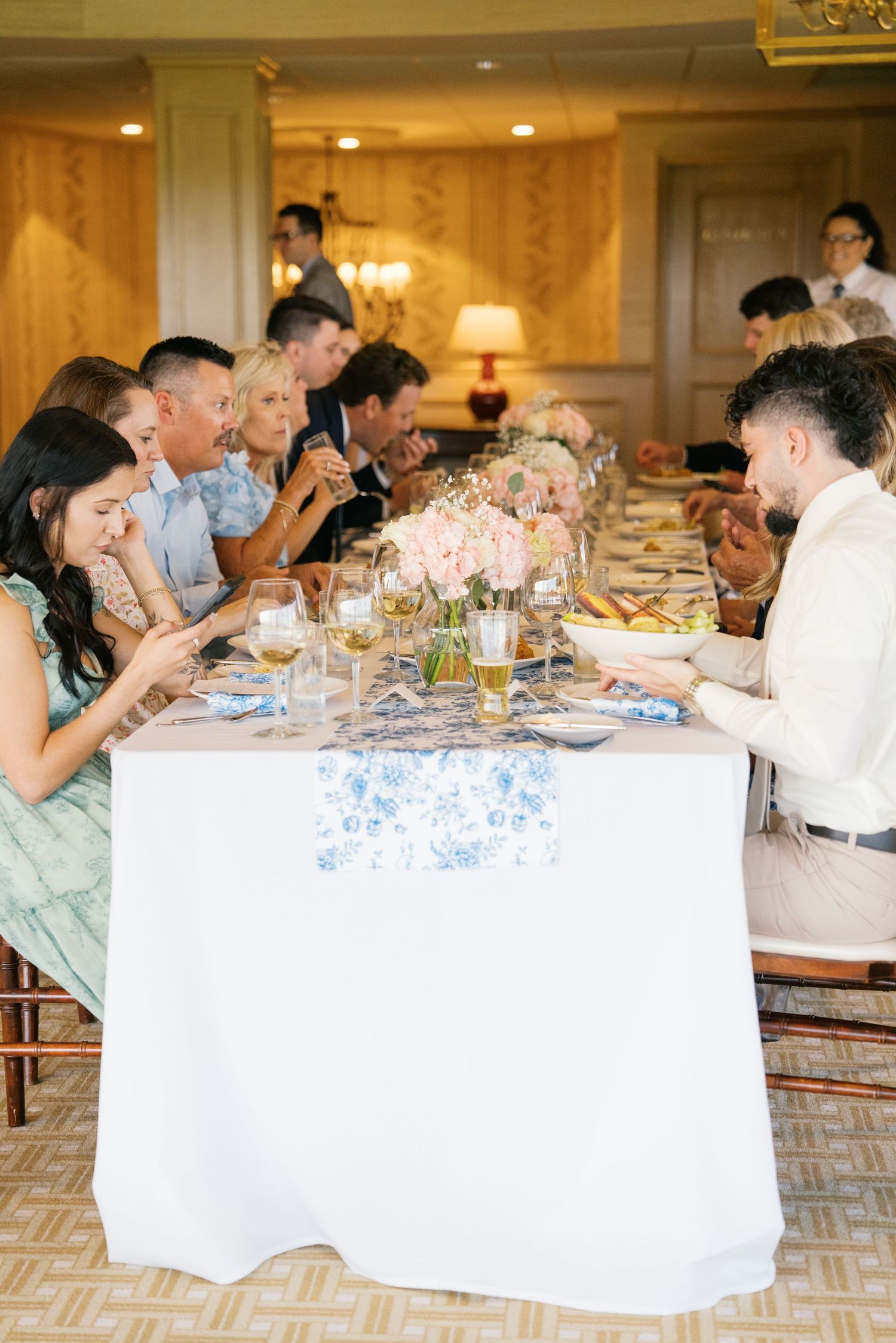 Elegant wedding reception dinner with guests seated at a long table decorated with pink floral centerpieces and white linens.